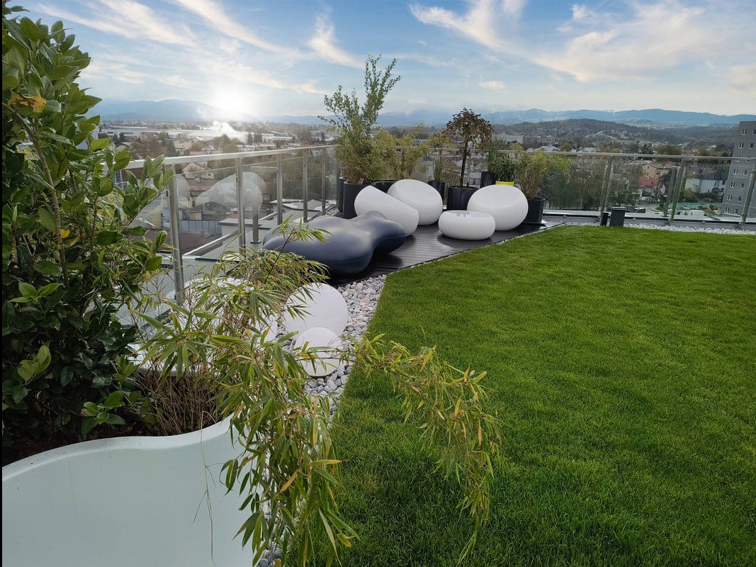 Nowoczesny taras na dachu z zielonym trawnikiem, białymi donicami i designerskimi meblami ogrodowymi na tle panoramy miasta i gór w oddali.