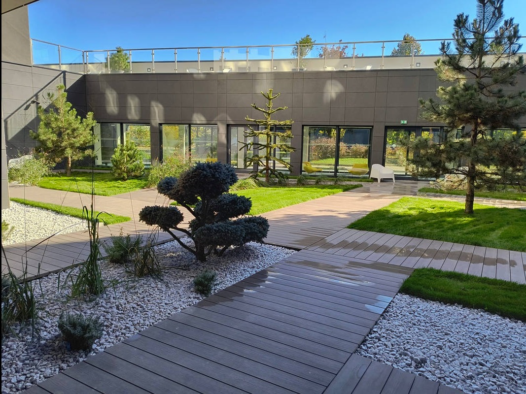 Nowoczesny ogród z drewnianymi ścieżkami, białym żwirem i ozdobnymi drzewami na tle budynku z dużymi oknami. Minimalistyczny design z elementami zieleni.