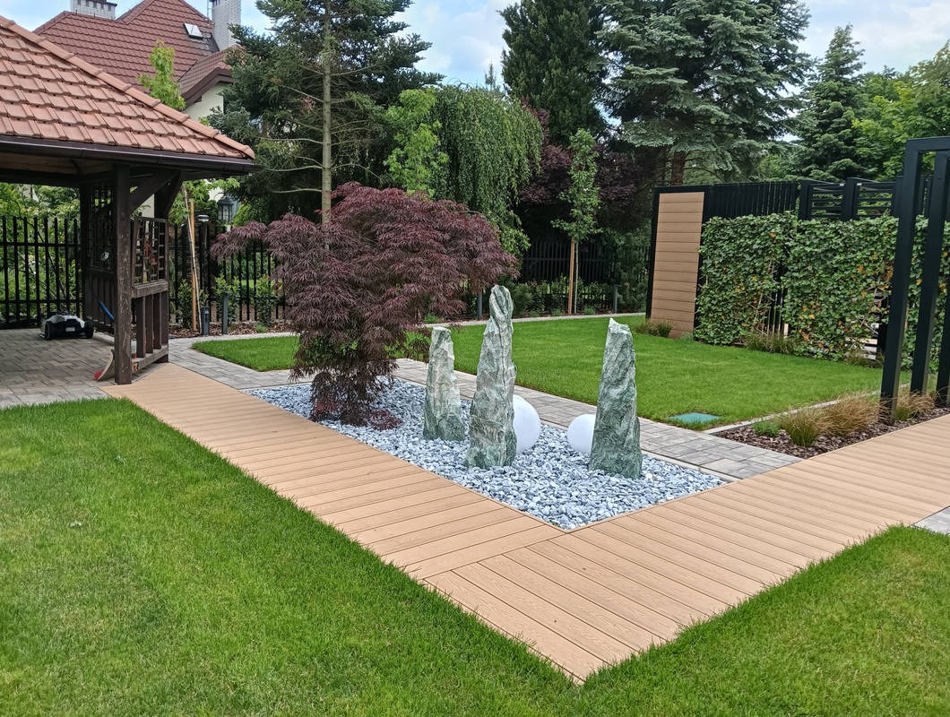 Urokliwy ogród z drewnianym tarasem, dekoracyjnym żwirem, rzeźbami z kamienia i starannie przystrzyżonym trawnikiem, otoczony zielenią i altaną.