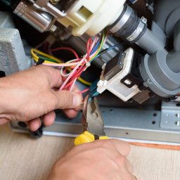 GTX SERVICE SP. Z O.O. SPÓŁKA KOMANDYTOWA - Naprawa zmywarki: elektryk przy pracy, używa szczypiec do przewodów wewnątrz urządzenia. Widoczne elementy pompy i okablowanie.