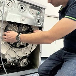 Naprawomat - Mężczyzna w trakcie naprawy pralki, skupiony na zakładaniu paska napędowego. Widoczny mechanizm pralki i narzędzia. Ujęcie z bliska, oświetlenie neutralne.