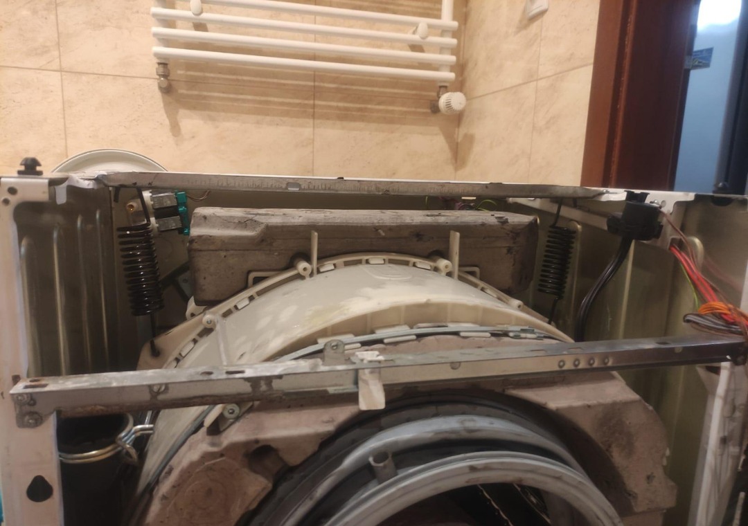 Wnętrze pralki automatycznej w trakcie naprawy, widoczne elementy konstrukcyjne i sprężyny amortyzujące bęben, na tle łazienki z grzejnikiem drabinkowym.