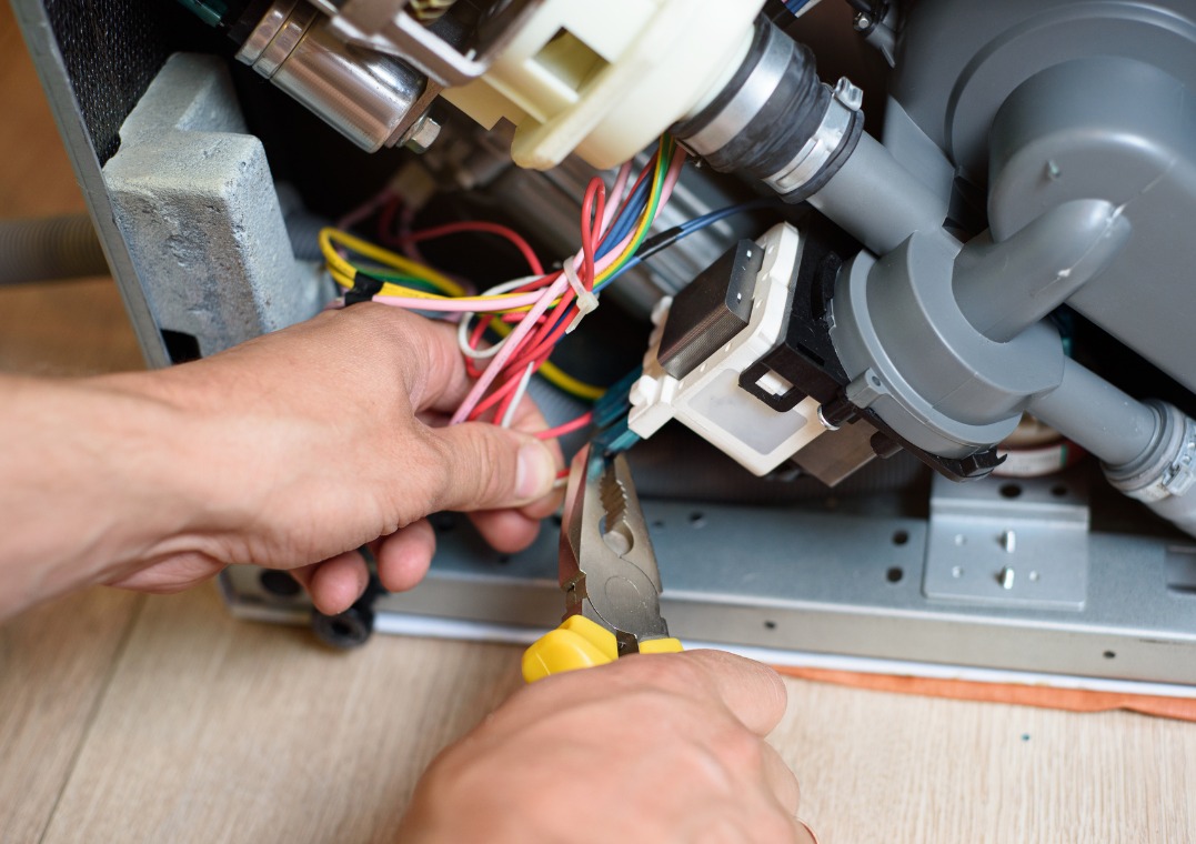 Naprawa zmywarki: elektryk przy pracy, używa szczypiec do przewodów wewnątrz urządzenia. Widoczne elementy pompy i okablowanie.