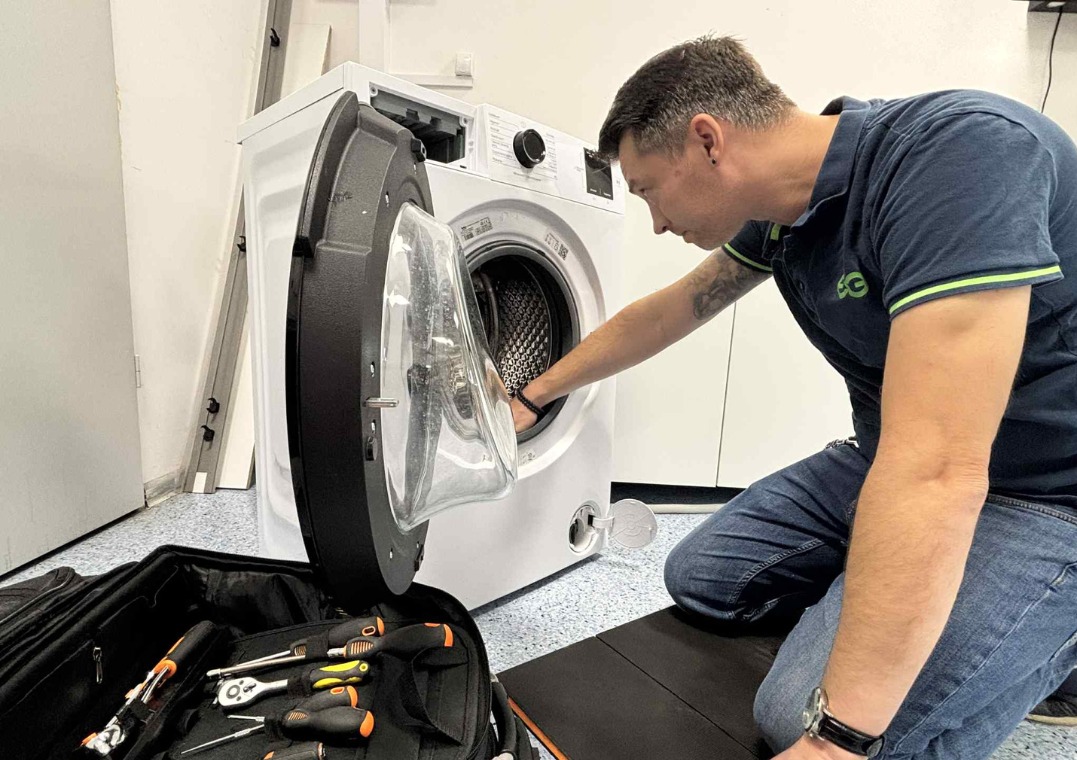 Serwisant w trakcie naprawy pralki z otwartymi drzwiczkami, obok rozłożona torba z narzędziami. Widoczny fragment podłogi i ściany.