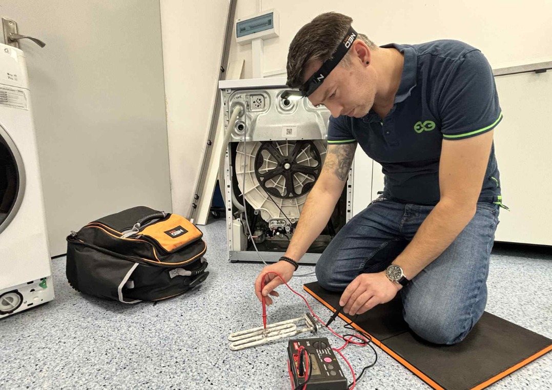 Mężczyzna w niebieskiej koszulce, klęczy przed otwartą pralką, mierząc grzałkę multimetrem. Obok torba narzędziowa NEO i druga pralka.