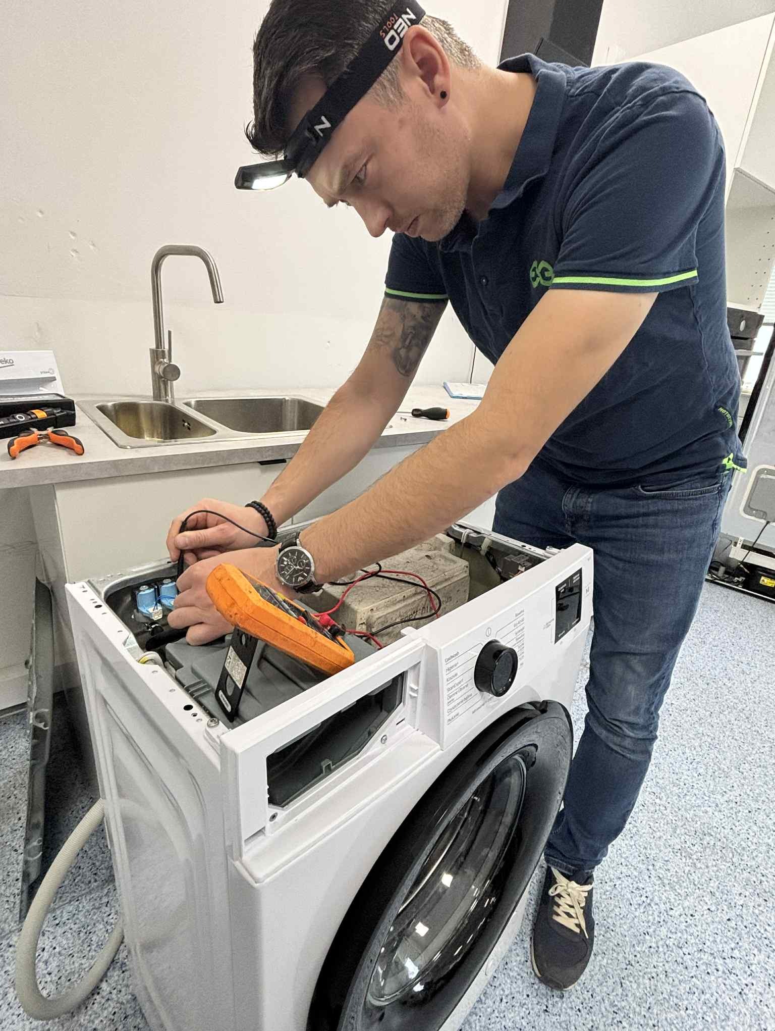 Mężczyzna w niebieskiej koszulce polo, z lampą czołową, naprawia pralkę za pomocą multimetru. Widoczne narzędzia i zlew w tle. Ujęcie z bliska, skupione na naprawie.