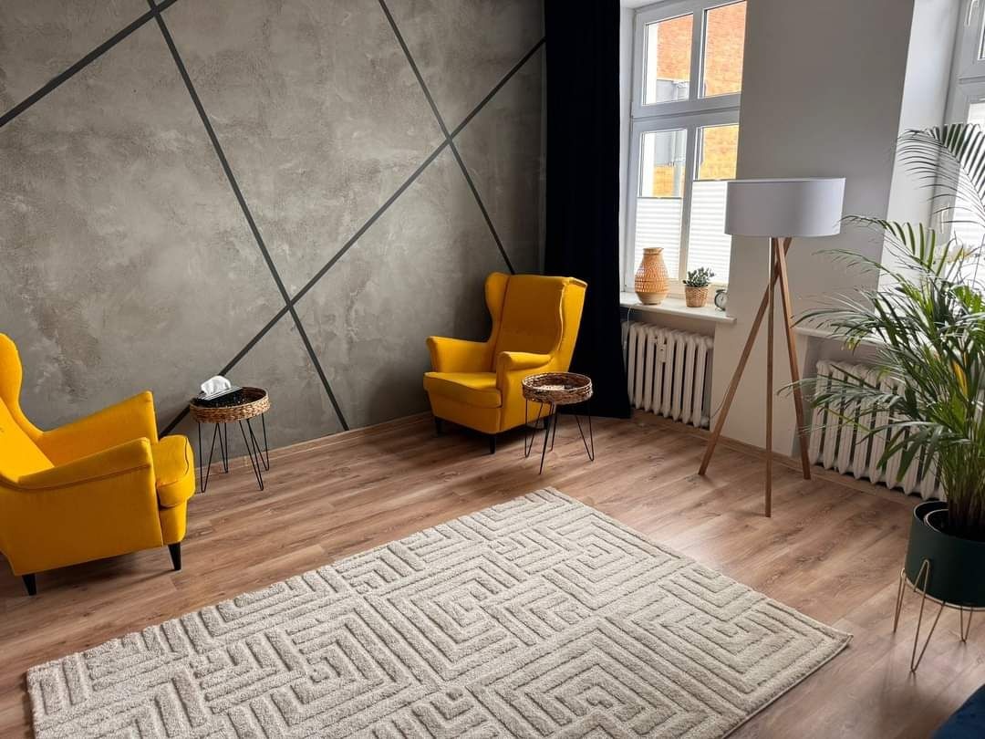 Jasne wnętrze gabinetu z dwoma żółtymi fotelami uszakami, stolikami, dywanem w geometryczne wzory i lampą podłogową. Na ścianie dekoracyjny beton z czarnymi liniami.
