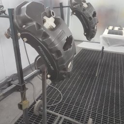 ZAKŁAD USŁUGOWO-HANDLOWO-PRODUKCYJNY Henryk Kasprzak AUTO SERVICE - Dwa zaciski hamulcowe Porsche po lakierowaniu, zawieszone na stojaku w kabinie lakierniczej. Widoczny napis 'PORSCHE' na zaciskach. Metalowa podłoga kratowa.