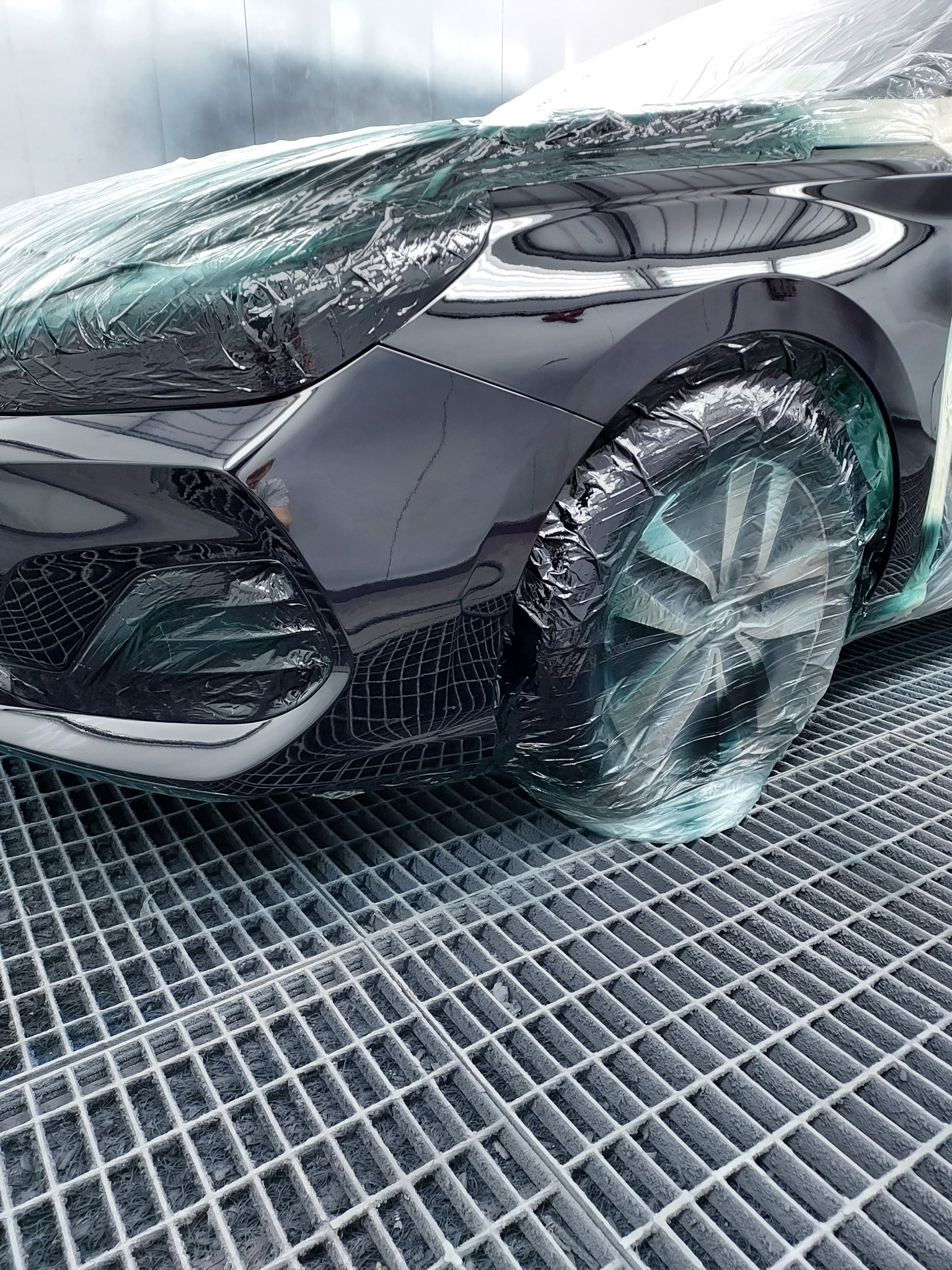 Częściowo zakryty folią, czarny samochód w kabinie lakierniczej, przygotowany do malowania. Widoczna kratka podłogowa.