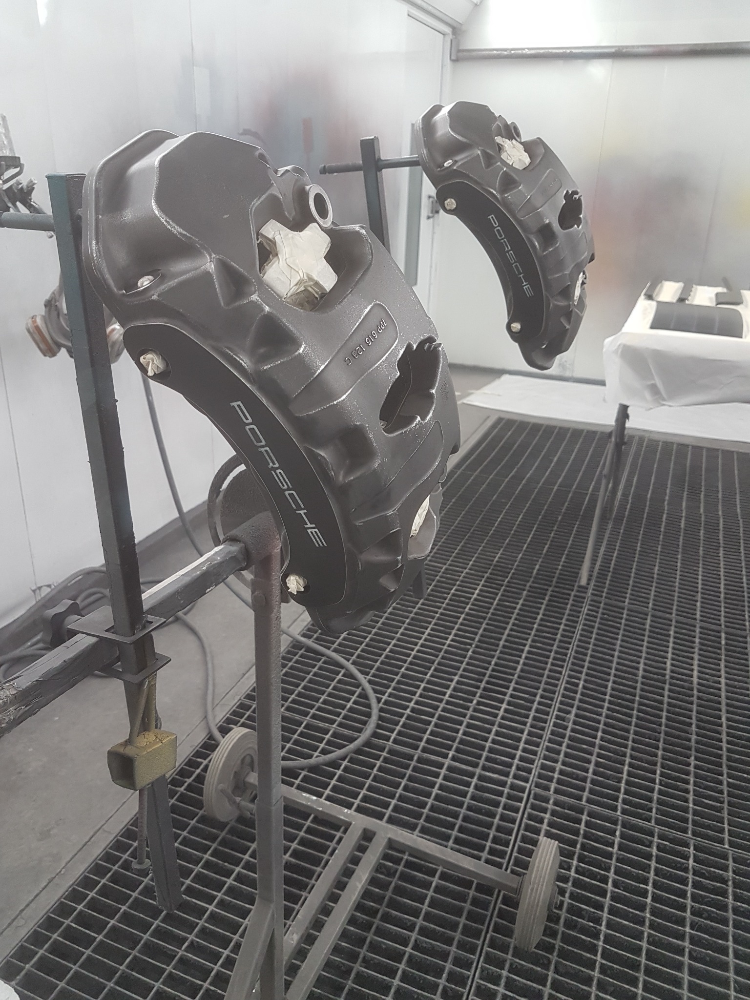 Dwa zaciski hamulcowe Porsche po lakierowaniu, zawieszone na stojaku w kabinie lakierniczej. Widoczny napis 'PORSCHE' na zaciskach. Metalowa podłoga kratowa.