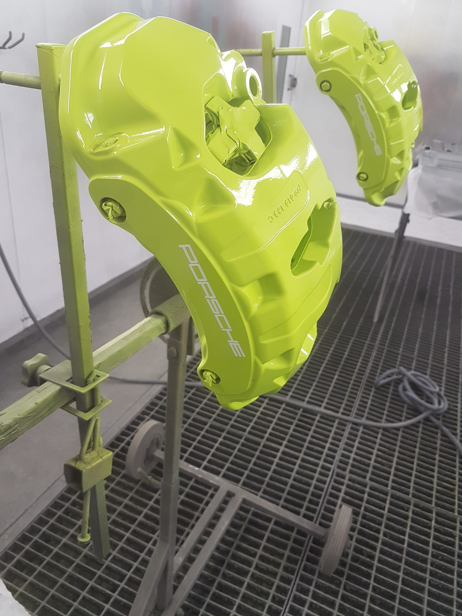 Pomalowane na jaskrawozielono zaciski hamulcowe Porsche, zawieszone na stojaku w lakierni. Widoczny napis Porsche na zacisku.