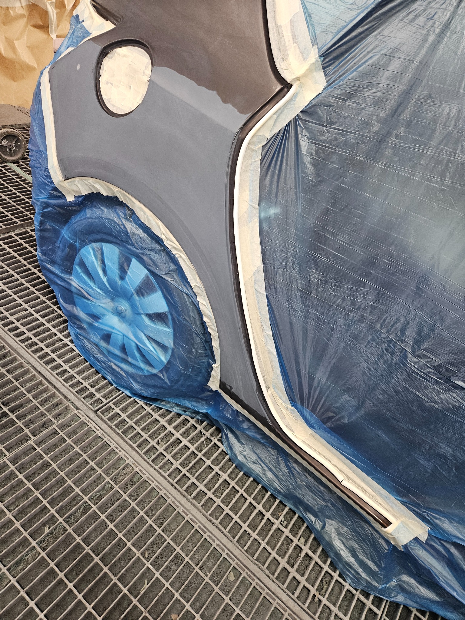 Przygotowanie auta do lakierowania: oklejone taśmą i folią elementy karoserii, widoczne koło w niebieskiej folii ochronnej, kratka podłogowa lakierni.