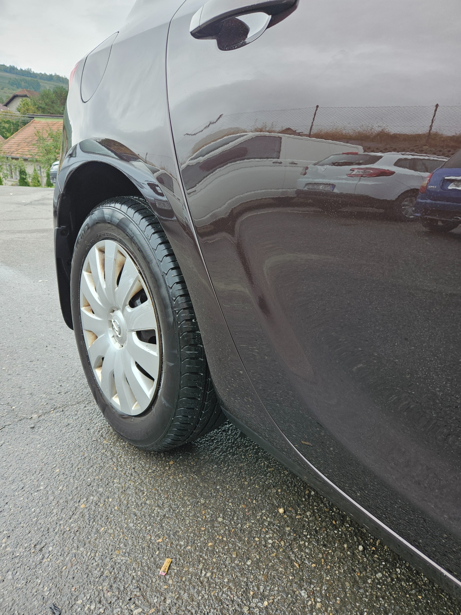 Błyszczący, brązowy bok auta z nową oponą i felgą. W lakierze odbijają się inne samochody. Mokry asfalt i niedopałek papierosa na ziemi.