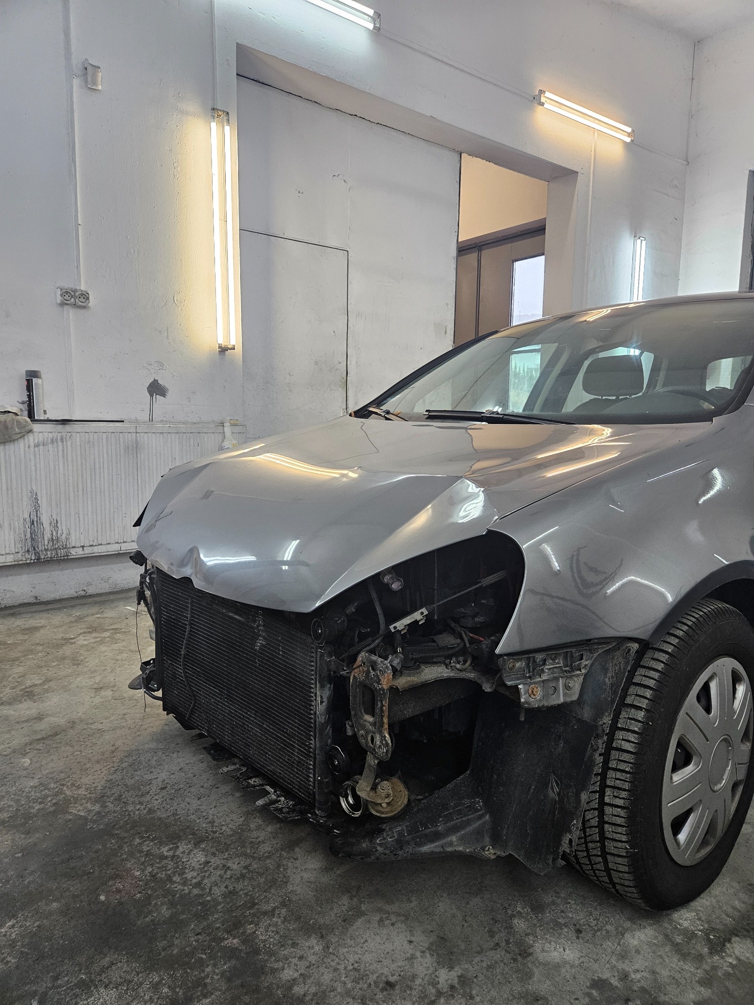 Uszkodzony przód srebrnego samochodu w warsztacie, widoczna zgnieciona maska i odsłonięty chłodnica. Oświetlenie jarzeniowe, betonowa podłoga.