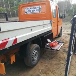 Pomarańczowy samoch&oacute;d dostawczy firmy montującej ogrodzenia, z otwartą skrzynką narzędziową, na tle lasu i częściowo zamontowanego ogrodzenia panelowego.