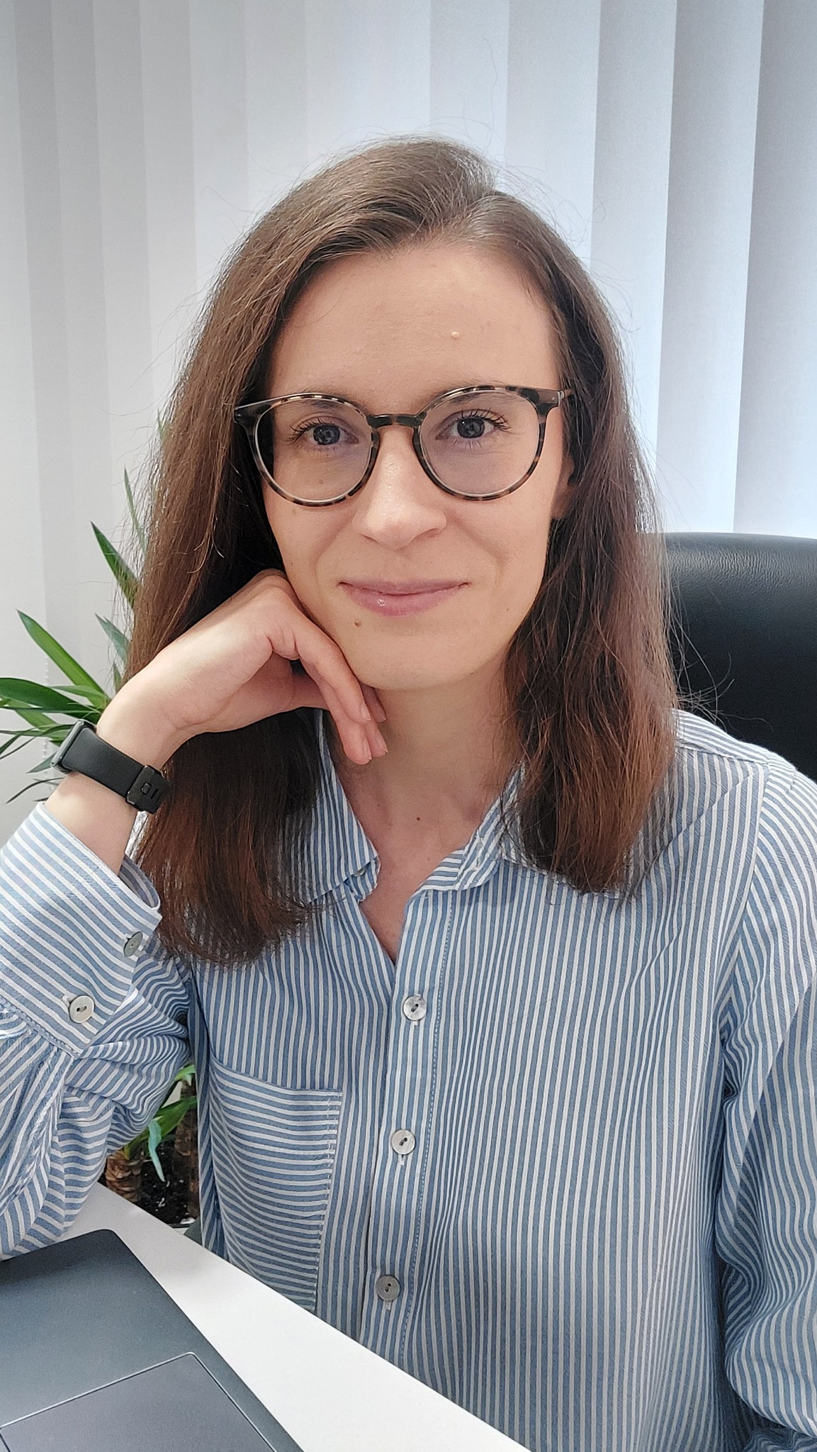 Portret uśmiechniętej kobiety w okularach i koszuli w paski, siedzącej przy biurku z laptopem i rośliną w tle, sugerujący profesjonalne konsultacje.