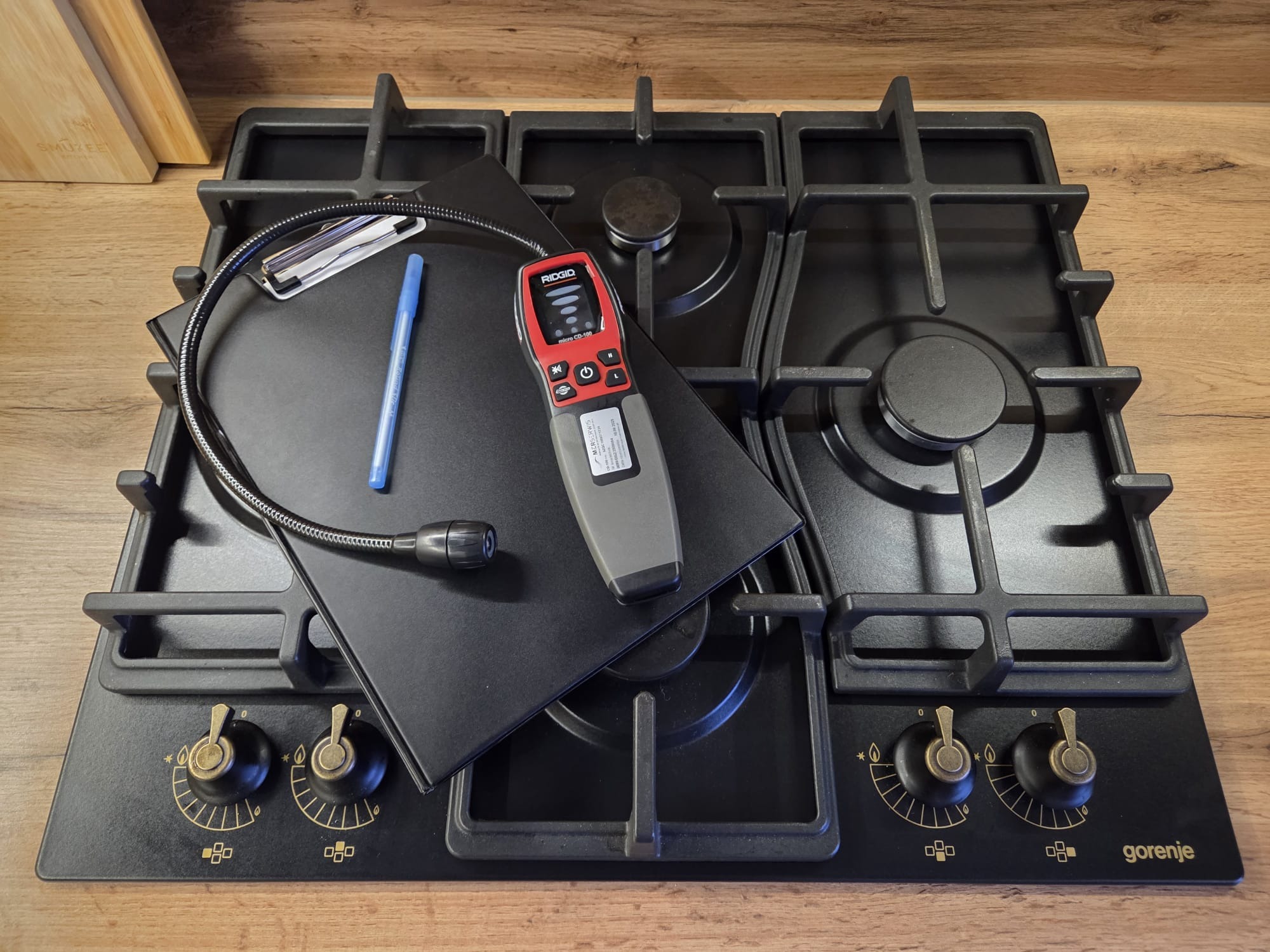 Czarna płyta gazowa Gorenje z detektorem nieszczelności gazu Ridgid, notatnikiem i długopisem, gotowa do sprawdzenia szczelności instalacji.