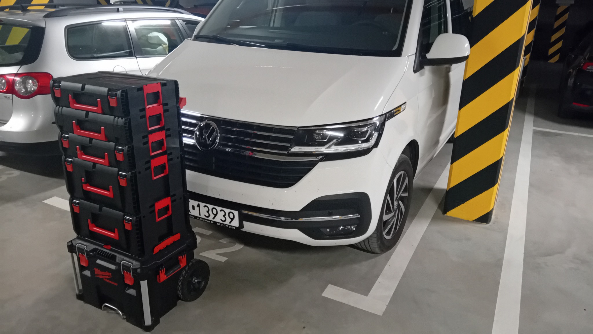 Zestaw skrzynek narzędziowych Milwaukee Packout na kółkach, obok białego samochodu VW Transporter na parkingu podziemnym. Widoczny słup z żółto-czarnymi pasami.