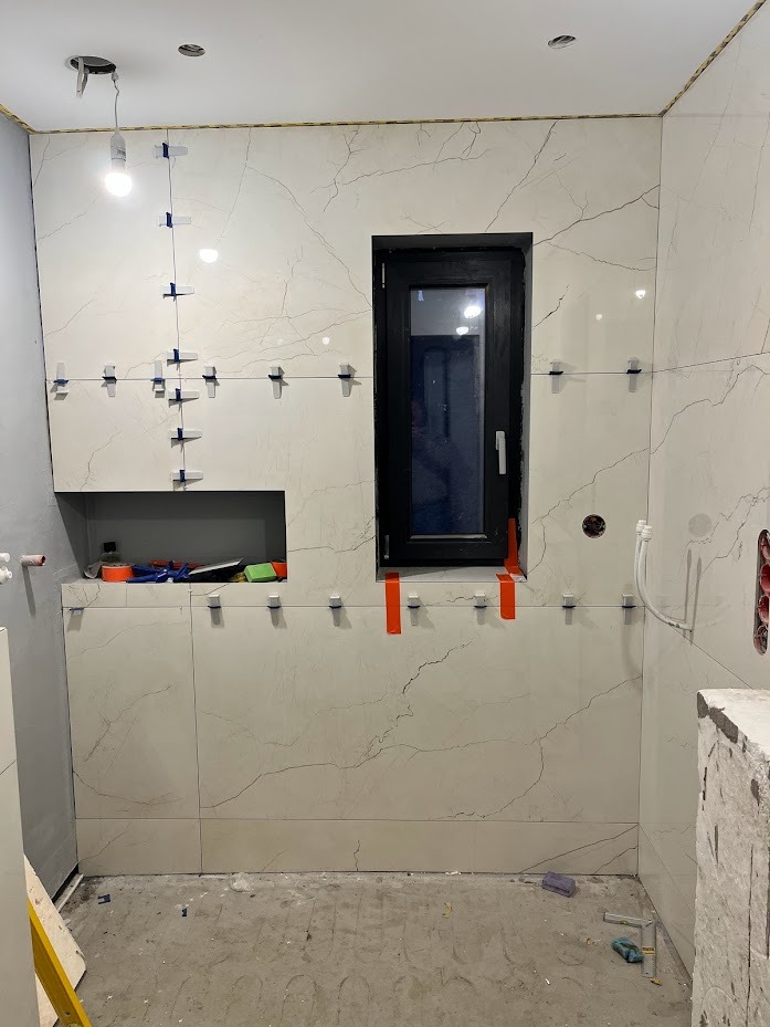 Ściana w trakcie układania płytek imitujących marmur, z użyciem krzyżyków dystansowych i poziomicy. Widoczne okno i wnęka. Prace glazurnicze w łazience.