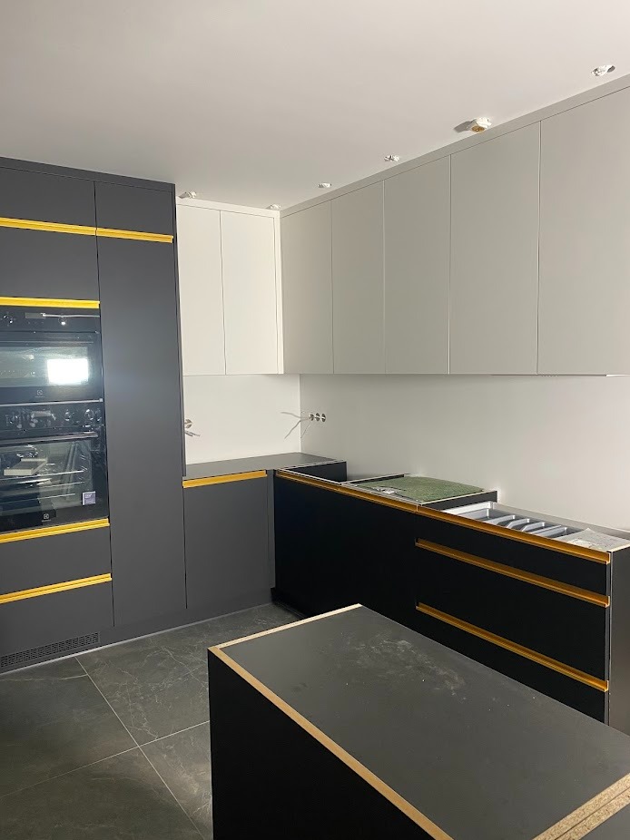 Częściowo zmontowana, nowoczesna kuchnia z czarnymi szafkami i złotymi akcentami, widoczne elementy AGD i szare płytki podłogowe. Etap montażu, surowy stan.