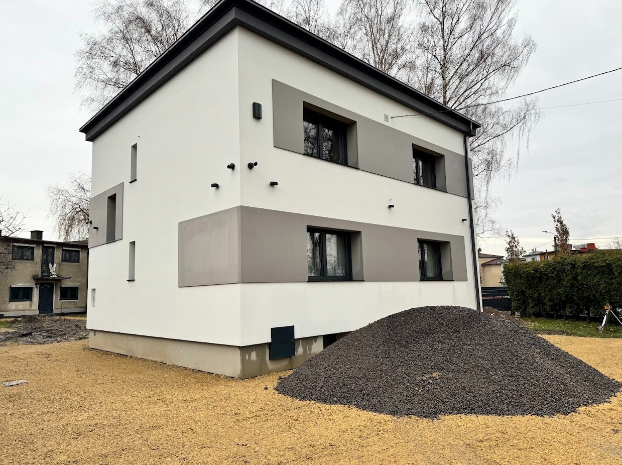 Dwukondygnacyjny dom z szarą elewacją i czarnymi detalami, otoczony żwirem. Na pierwszym planie sterta kamieni. Widok na budynek z boku, w tle drzewa.