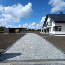 Roman Widła - Nowoczesny podjazd z szarej kostki brukowej prowadzący do białego domu z czarnym dachem, w tle błękitne niebo z chmurami i słup wysokiego napięcia.