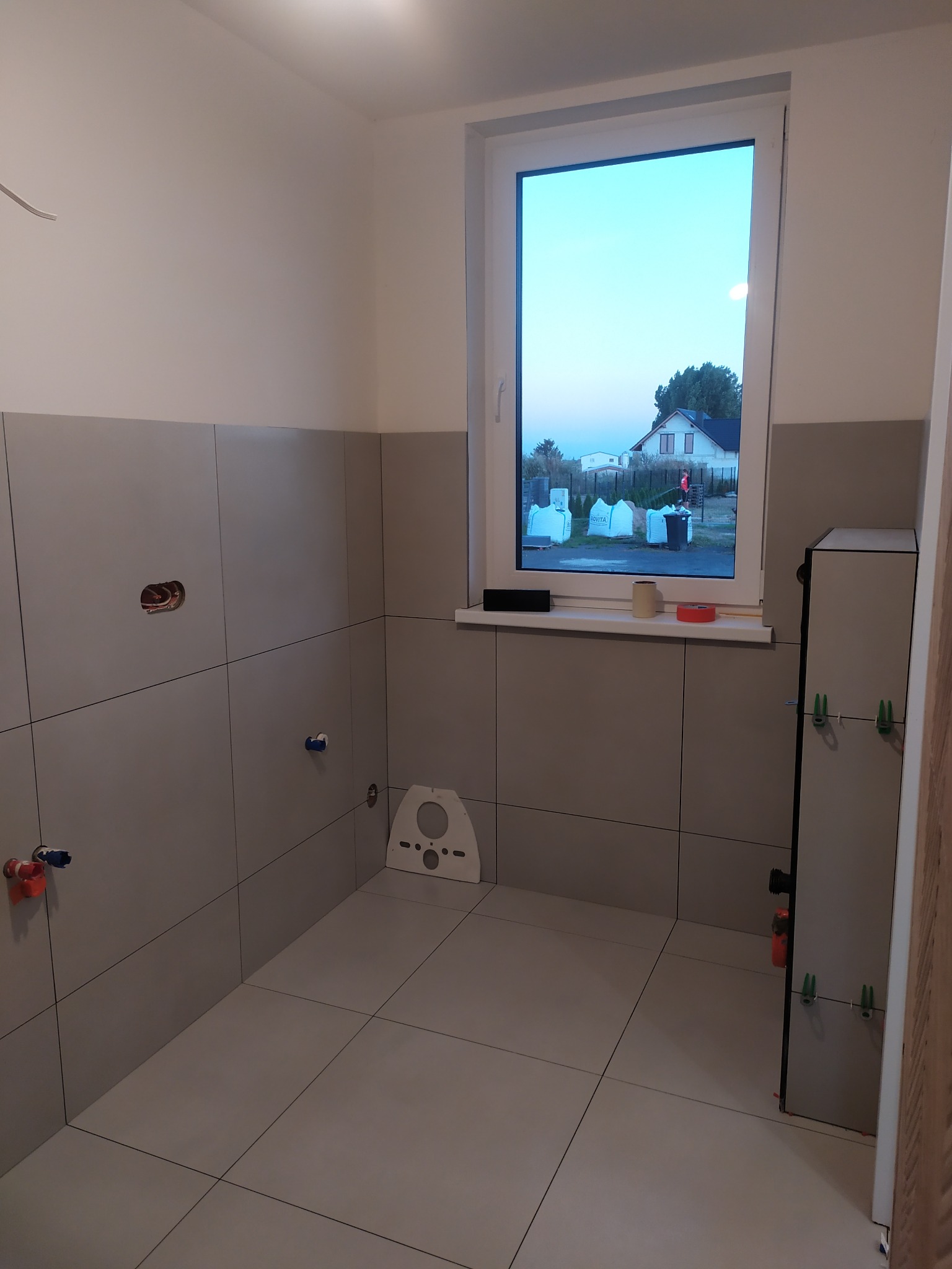 Wnętrze łazienki w trakcie remontu: szare płytki na ścianach do połowy wysokości, okno z widokiem na dom, przygotowane przyłącza wodne i kanalizacyjne.