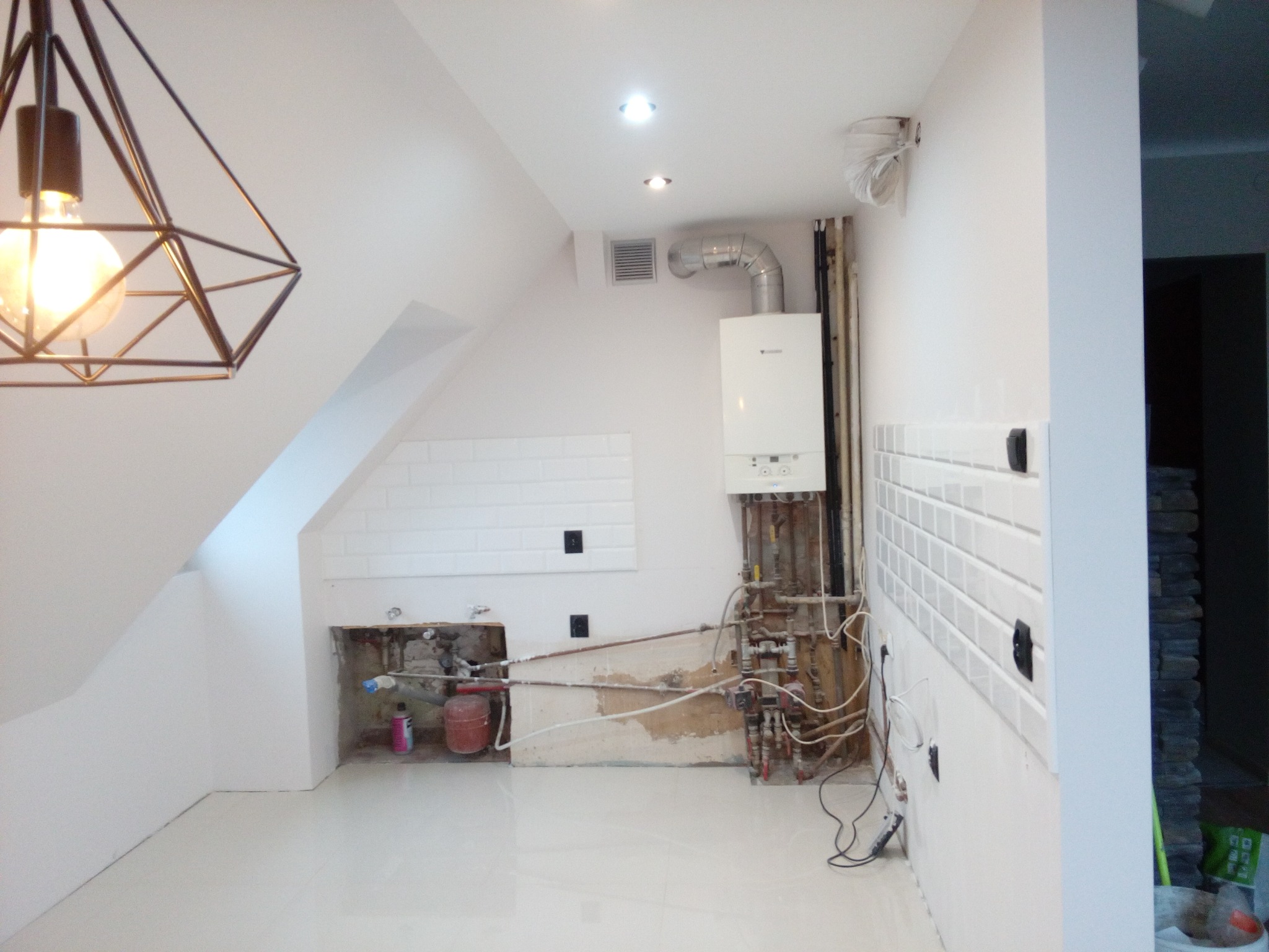 Wnętrze w trakcie remontu: białe ściany z fragmentami płytek, odsłonięte instalacje hydrauliczne i elektryczne, wiszący nowoczesny żyrandol. Widoczny piec gazowy.
