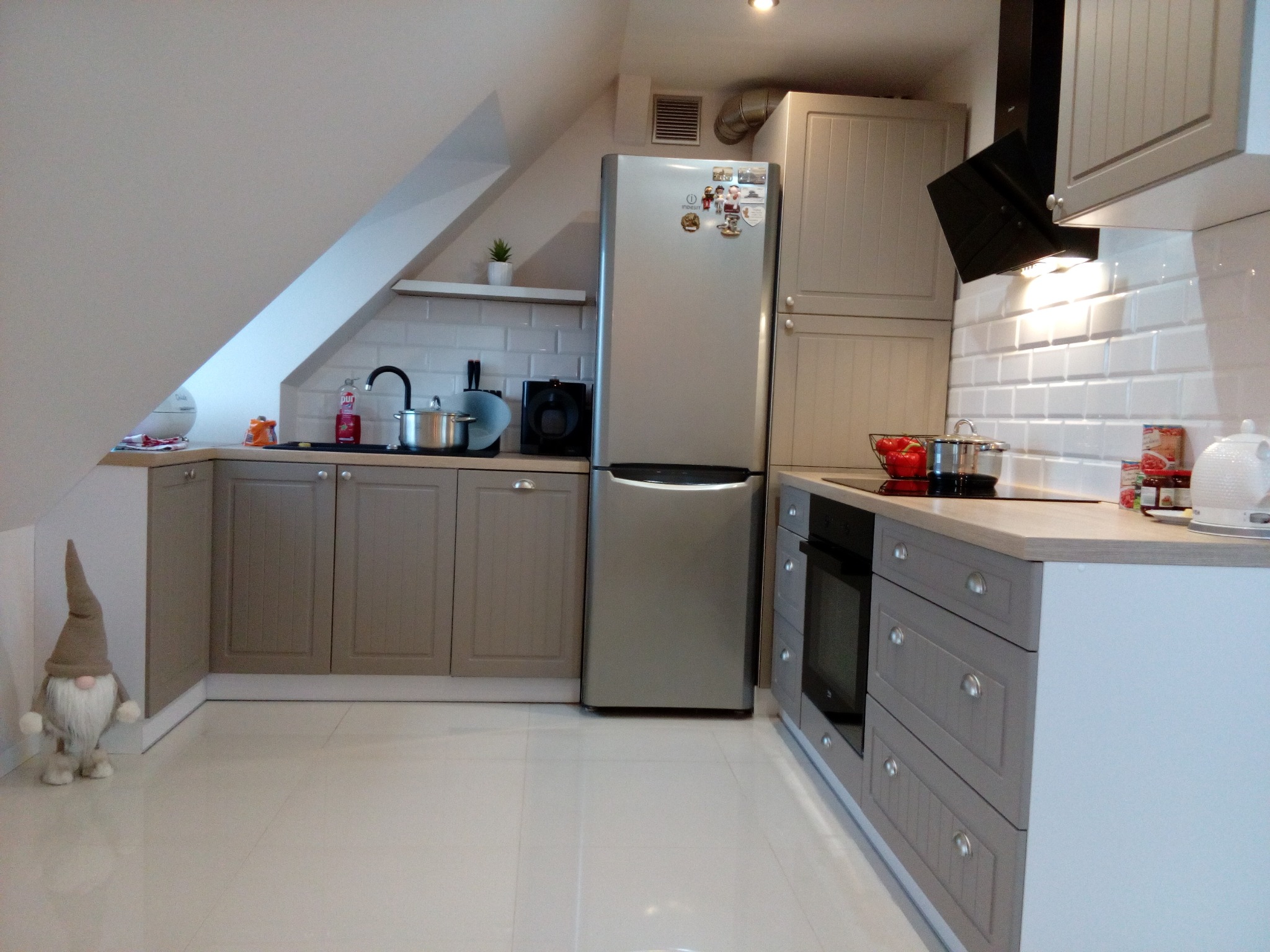 Jasna kuchnia na poddaszu z beżowymi szafkami, srebrną lodówką i białymi płytkami. Uwagę przykuwa dekoracyjny skrzat w rogu pomieszczenia.