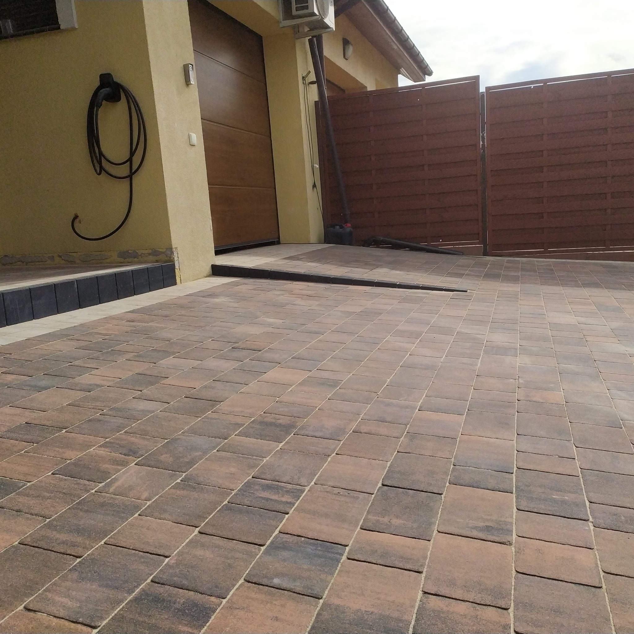 Ułożona kostka brukowa w odcieniach brązu na podjeździe, z widocznym fragmentem bramy garażowej i ogrodzenia w tle. Widoczny wąż na ścianie budynku.