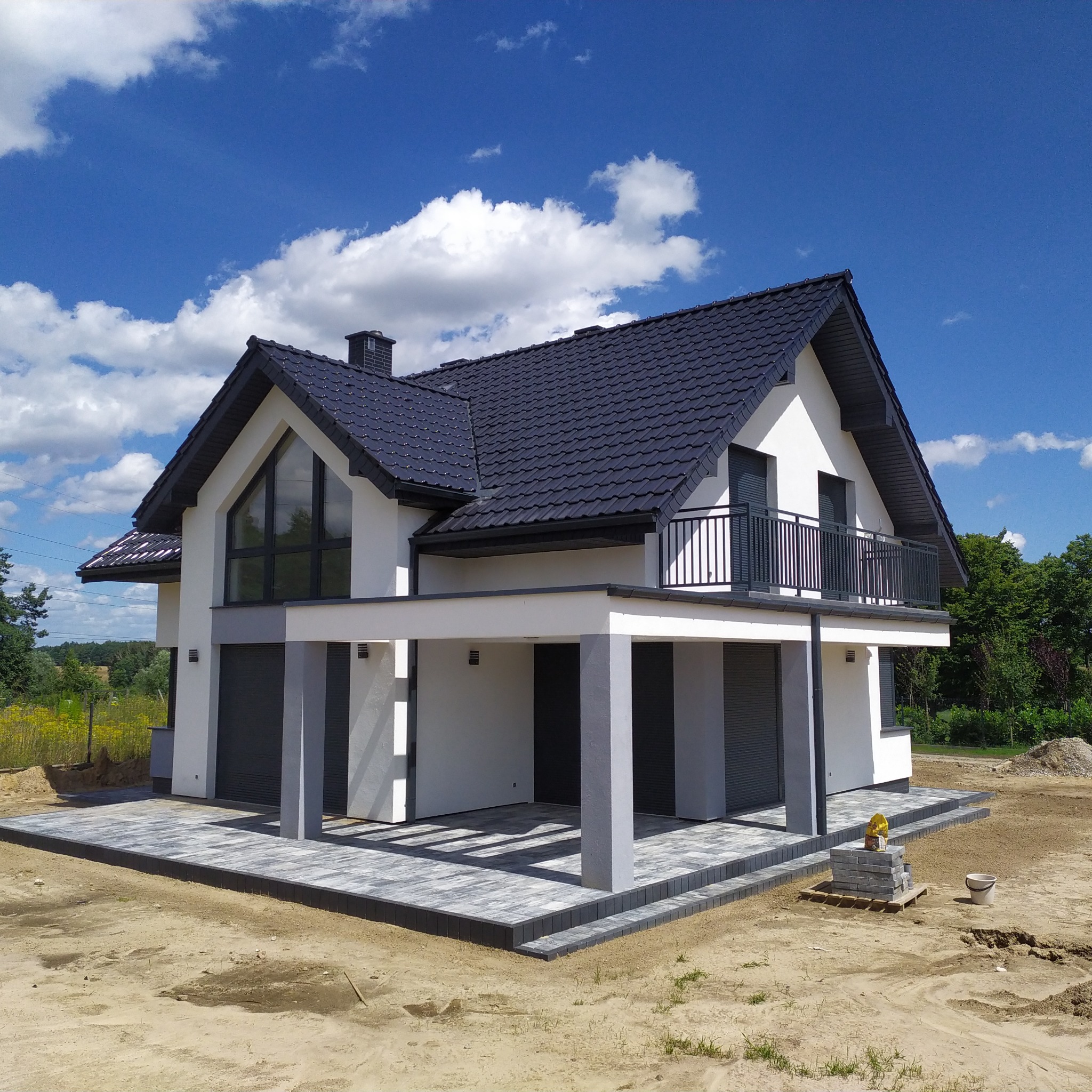 Nowoczesny dom jednorodzinny z szarą dachówką i wybrukowanym tarasem, widok z zewnątrz w słoneczny dzień. Na pierwszym planie kostka brukowa i przygotowany teren.