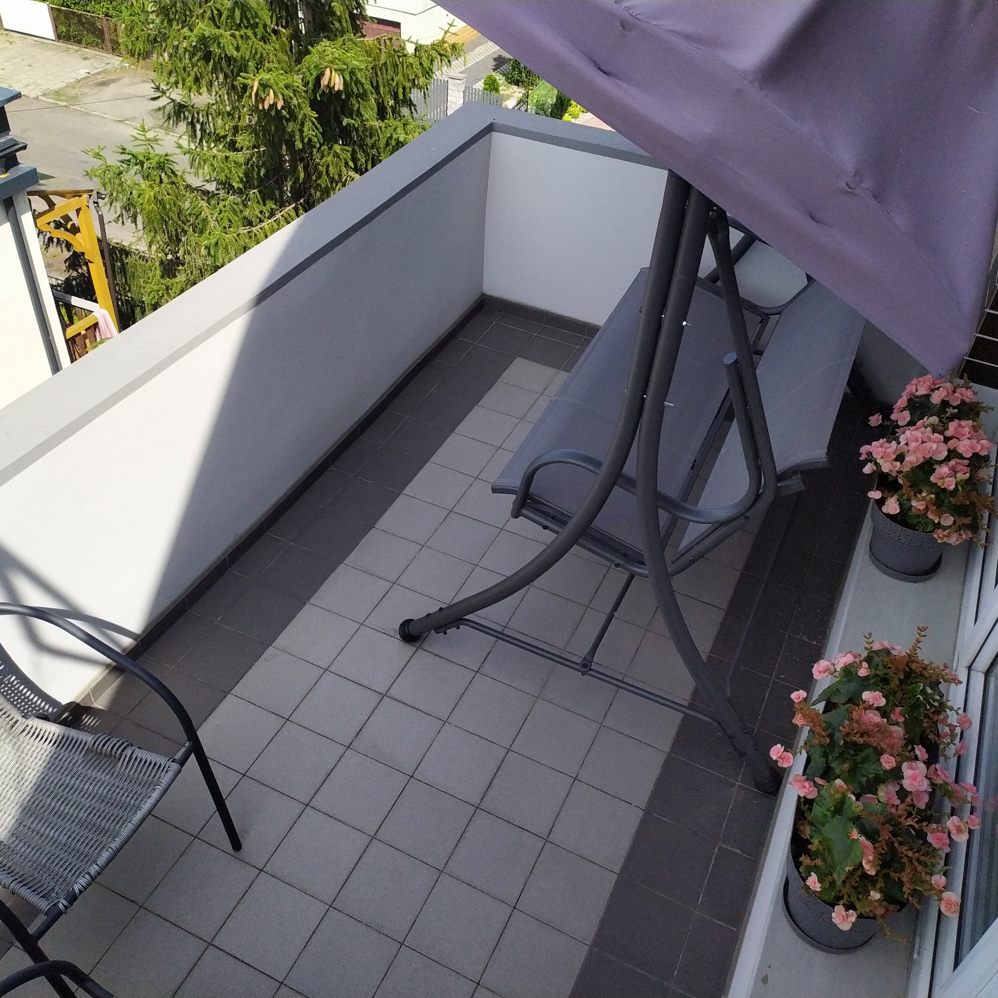 Umeblowany balkon z szarą huśtawką, krzesłem i kwiatami w donicach. Płytki podłogowe w odcieniach szarości. Widok z góry na drzewa za barierką.
