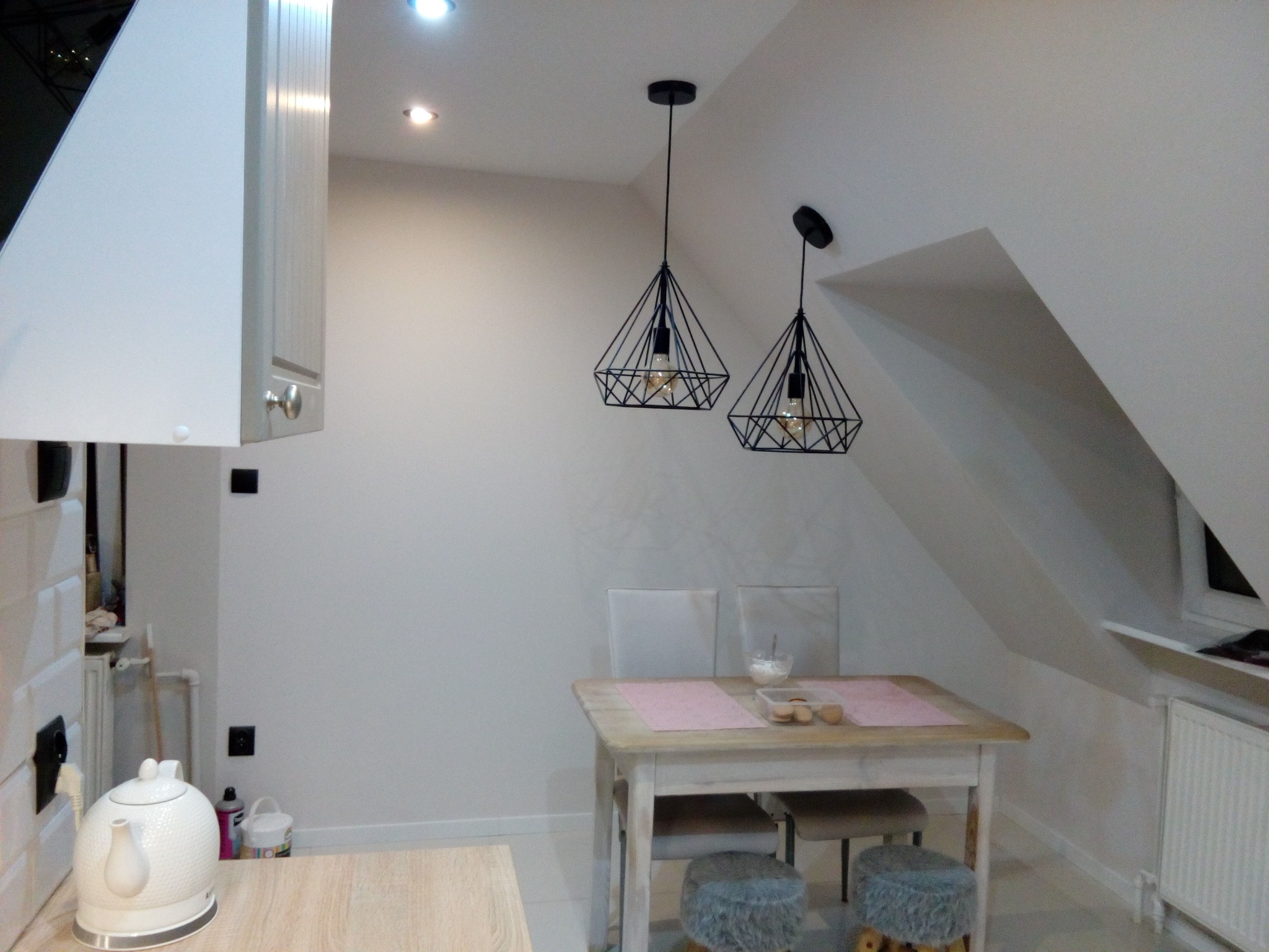 Jasna kuchnia na poddaszu z dwoma lampami w stylu loft, drewnianym stołem i białymi krzesłami. Widoczny fragment zabudowy kuchennej i czajnik elektryczny.