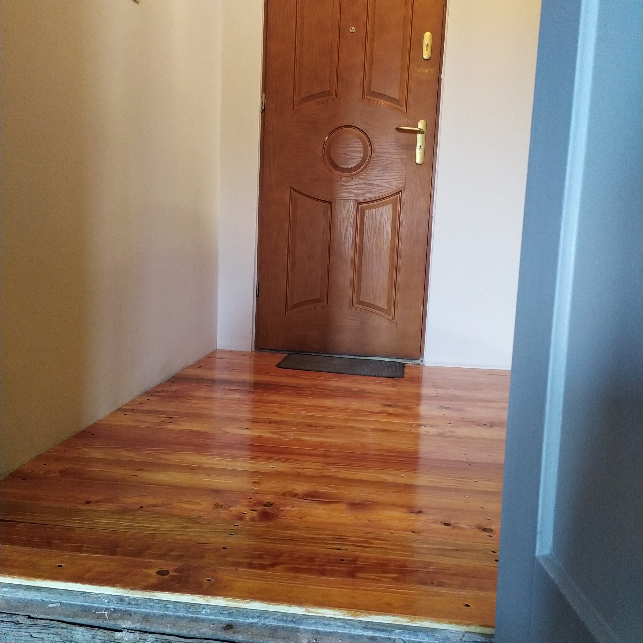 Wykończona podłoga z drewna w odcieniach brązu, widoczna przy wejściu do pomieszczenia z drewnianymi drzwiami i pomalowanymi ścianami. Widoczny fragment futryny.