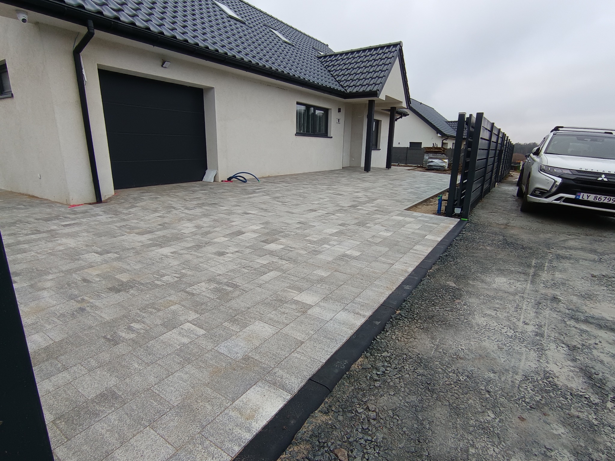 Ułożona kostka brukowa na podjeździe przed domem z czarną bramą garażową i nowoczesnym ogrodzeniem panelowym. Widoczny samochód osobowy.