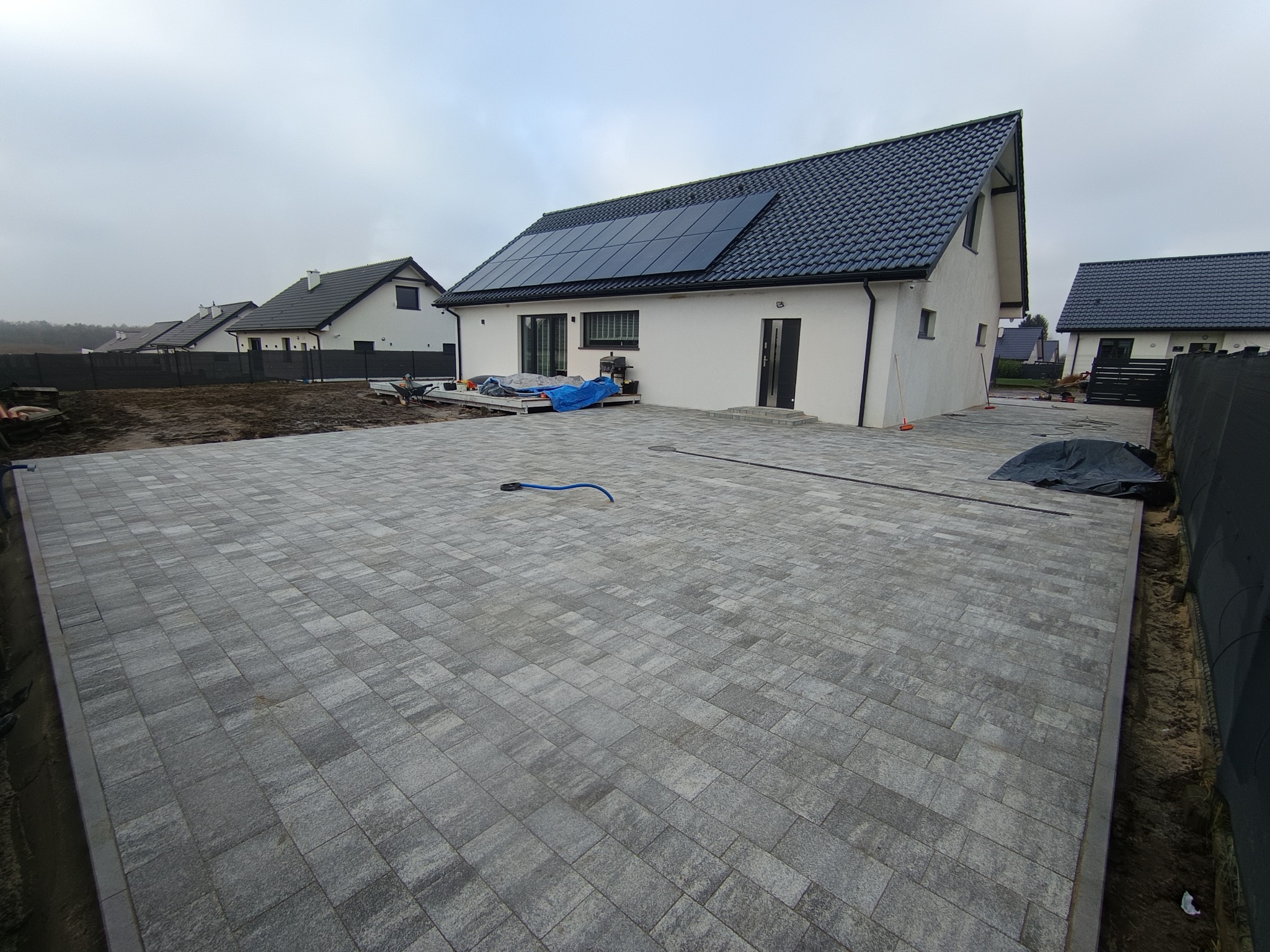 Ułożona szara kostka brukowa na podjeździe przed nowoczesnym domem z panelami słonecznymi. Widoczne sąsiednie domy i pochmurne niebo.