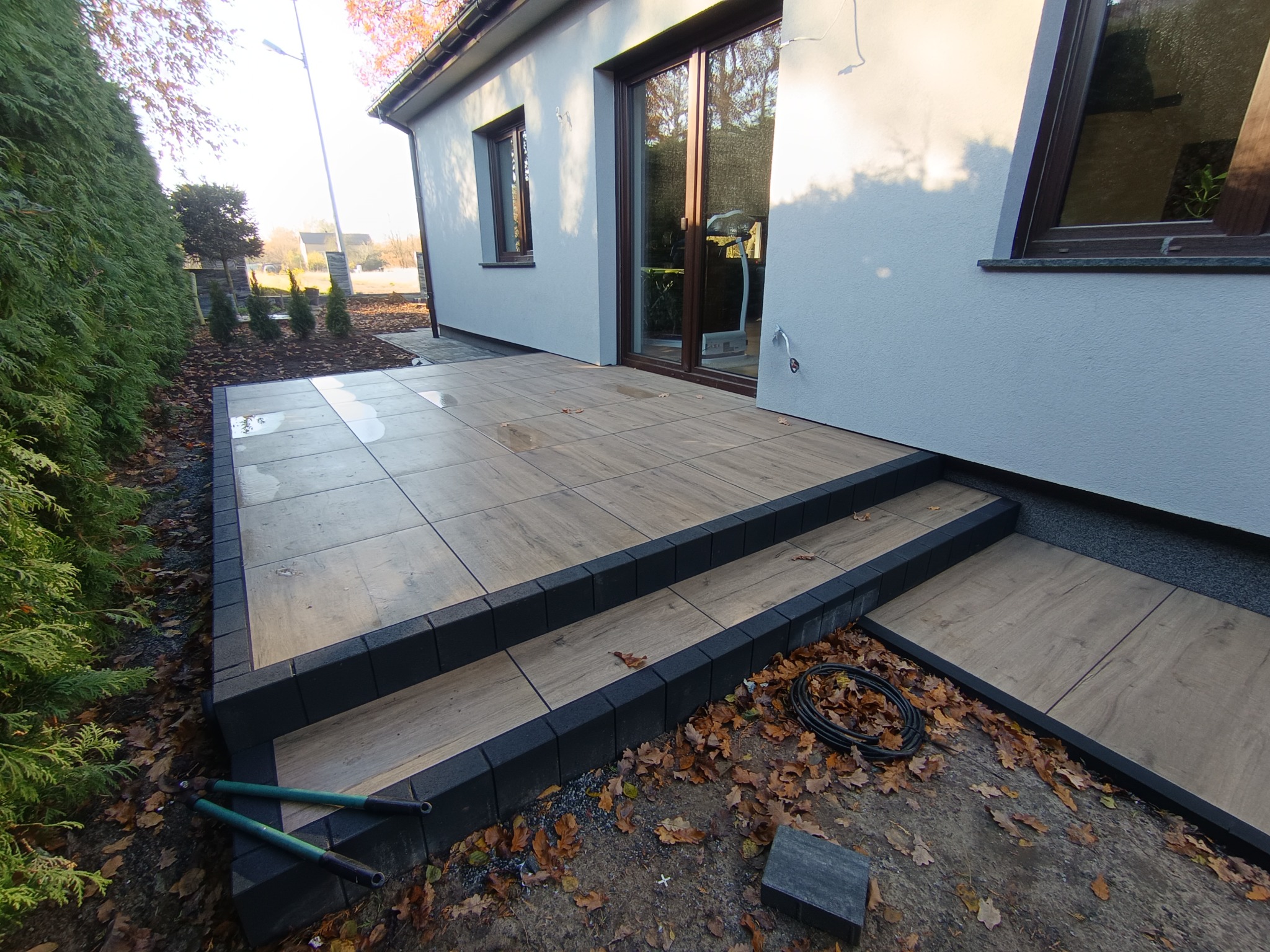 Wykończony taras z drewnopodobnych płytek i czarnych obrzeży przy domu, z widocznymi schodami i narzędziami ogrodniczymi. Jesienne liście na ziemi.