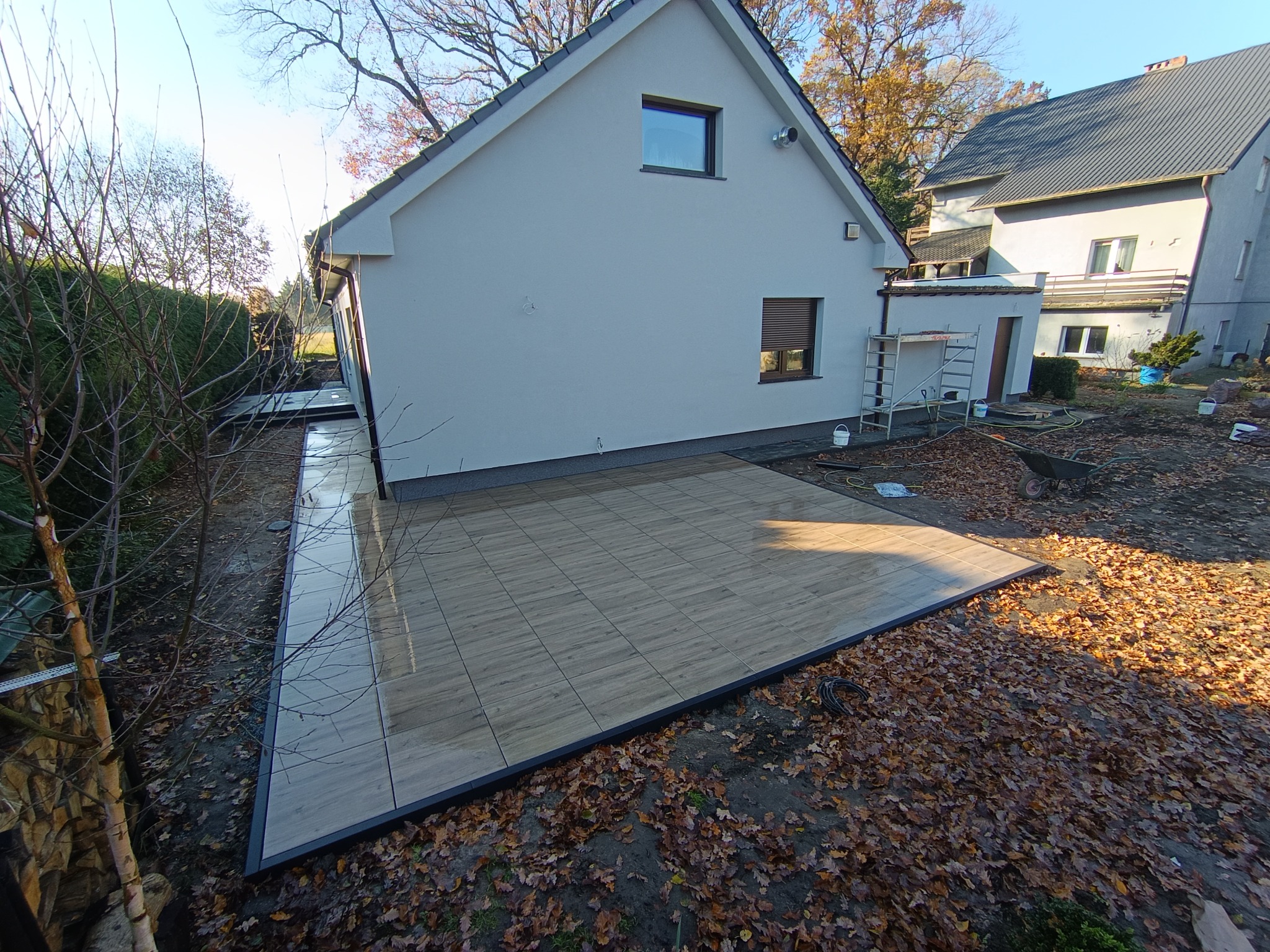 Nowoczesny taras z płytek drewnopodobnych, obrzeża w kolorze antracytu, przylegający do białego domu z szarym dachem. Jesienne liście na ziemi.