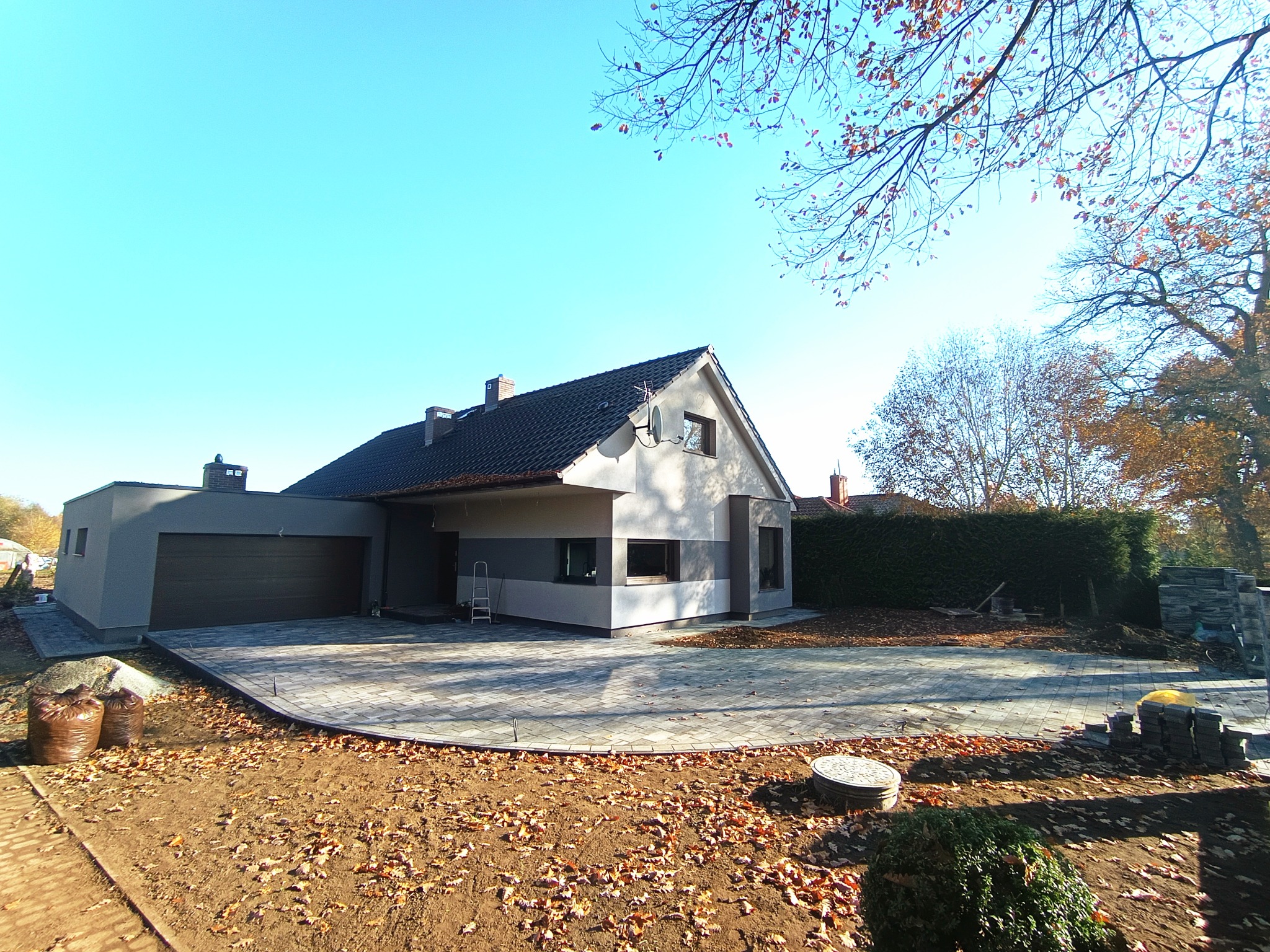 Nowoczesny dom z szarą elewacją i brukowanym podjazdem w jesiennej scenerii. Widoczne worki z materiałem budowlanym i ułożona kostka brukowa.