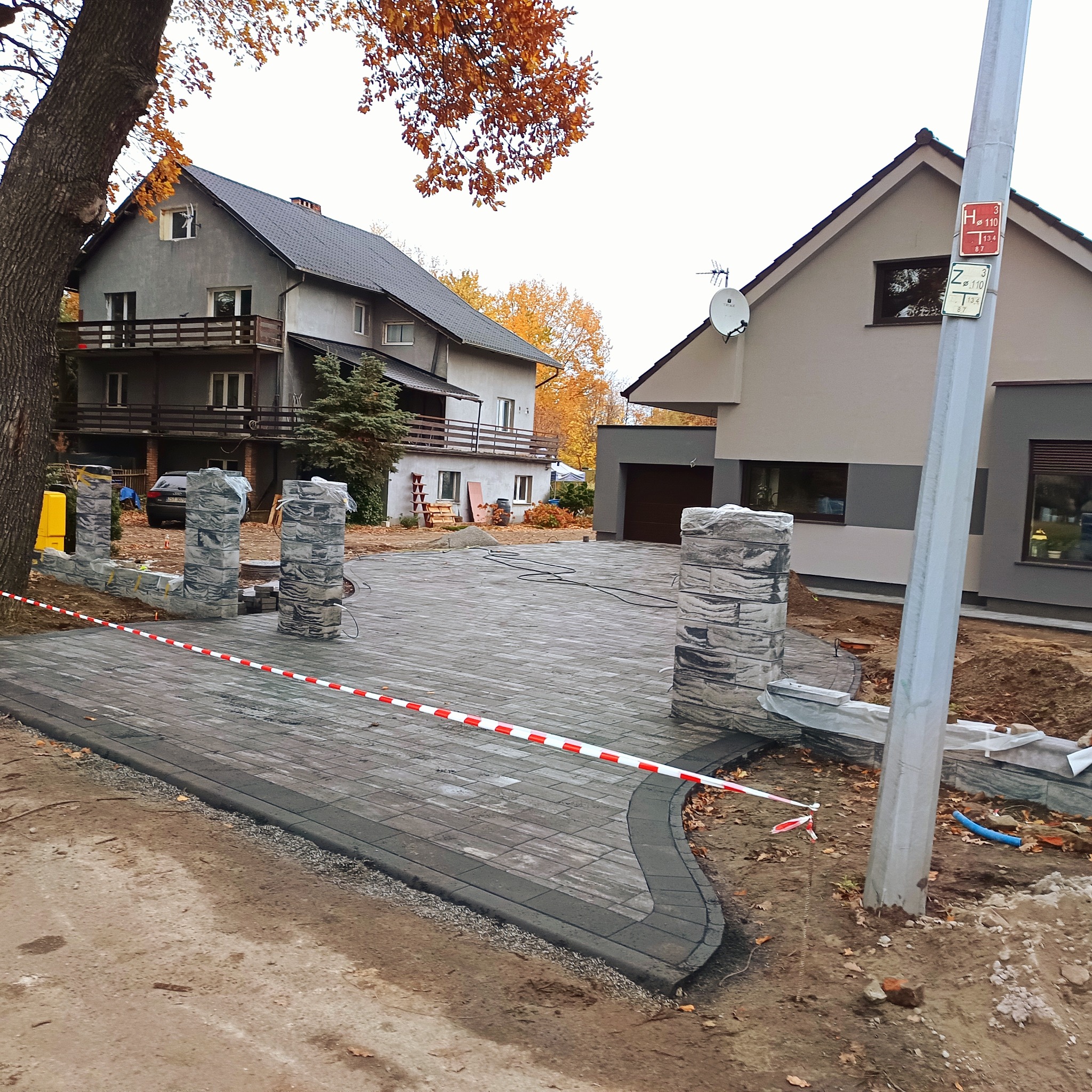 Układanie kostki brukowej na podjeździe do domu z elementami ogrodzenia z kamienia. Widoczne dwa budynki w tle i jesienne drzewa.