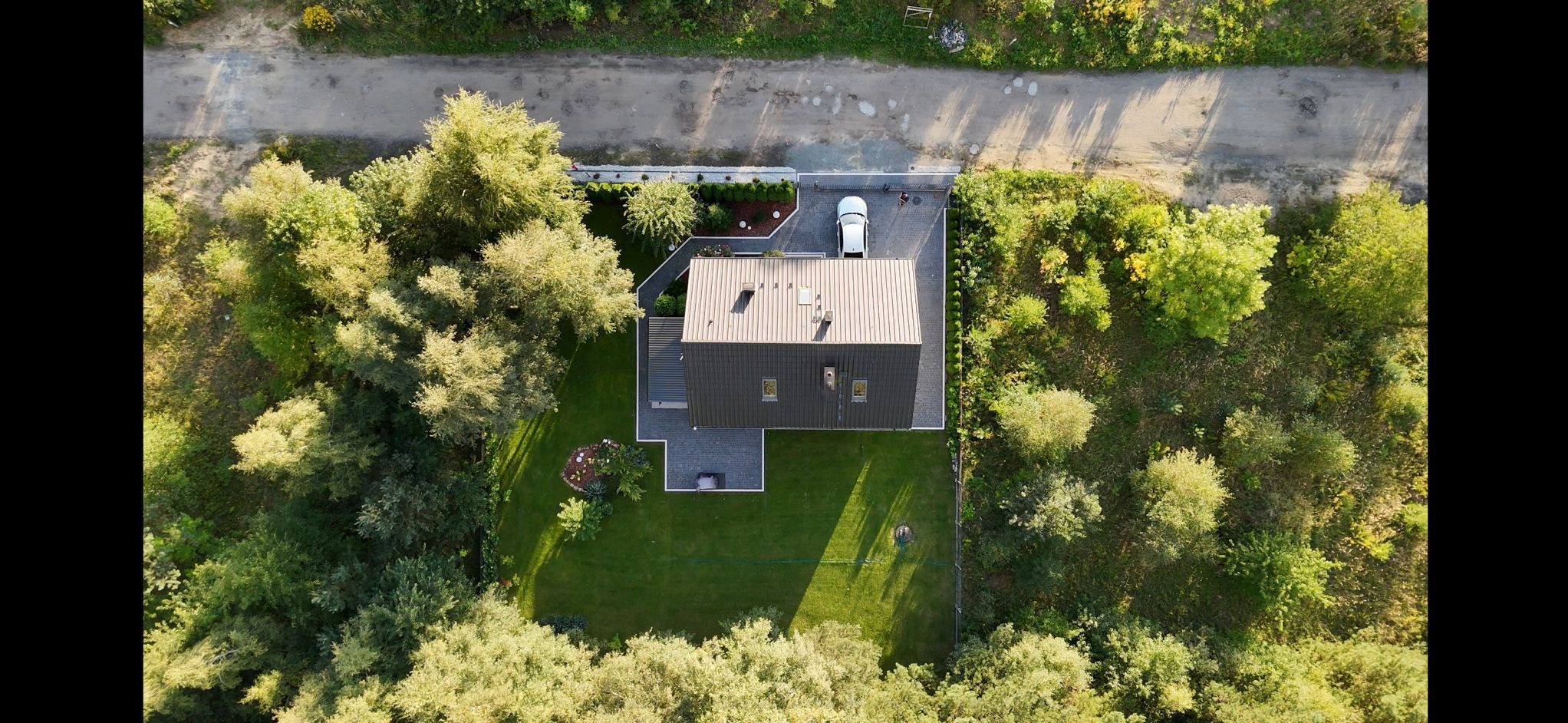 Zdjęcie z drona przedstawia dom z ciemnym dachem, zadbanym trawnikiem, ozdobnymi rabatami i podjazdem z kostki, na którym stoi biały samochód. Widoczna droga gruntowa i zieleń.