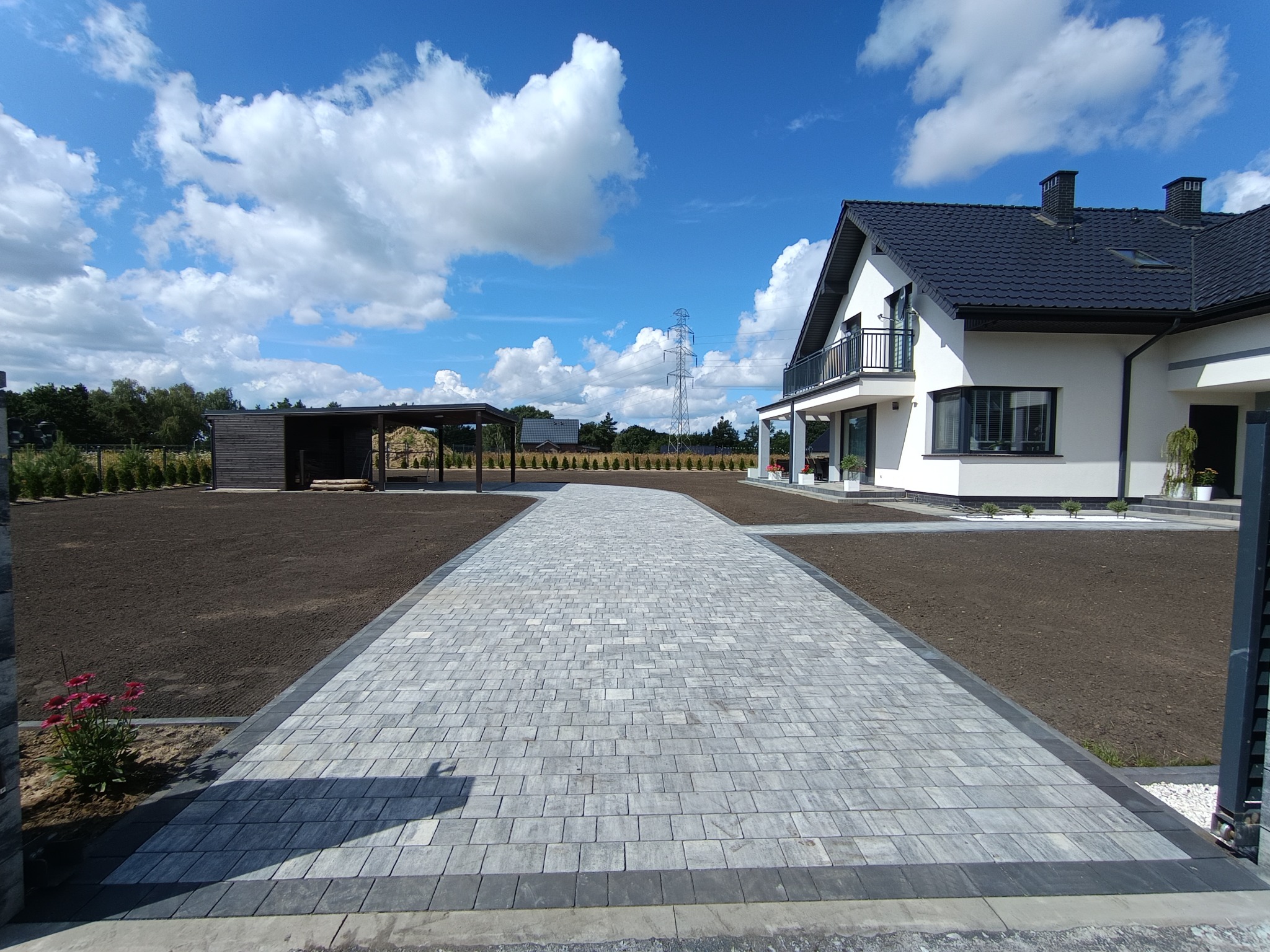 Nowoczesny podjazd z szarej kostki brukowej prowadzący do białego domu z czarnym dachem, w tle błękitne niebo z chmurami i słup wysokiego napięcia.
