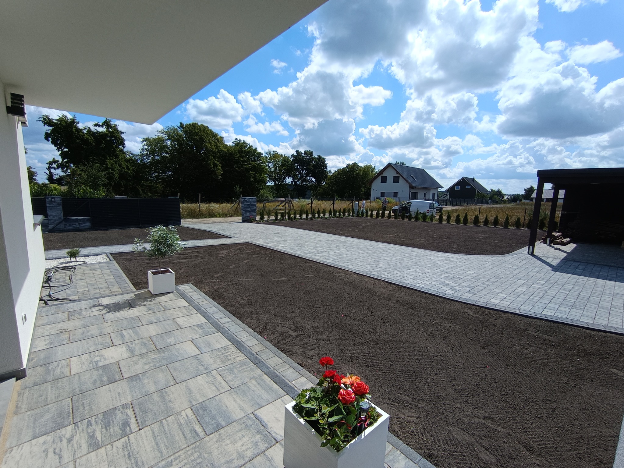 Nowoczesny podjazd z kostki brukowej w odcieniach szarości, świeżo obsadzony trawnik, minimalistyczna architektura domu i dekoracyjne rośliny w donicach.