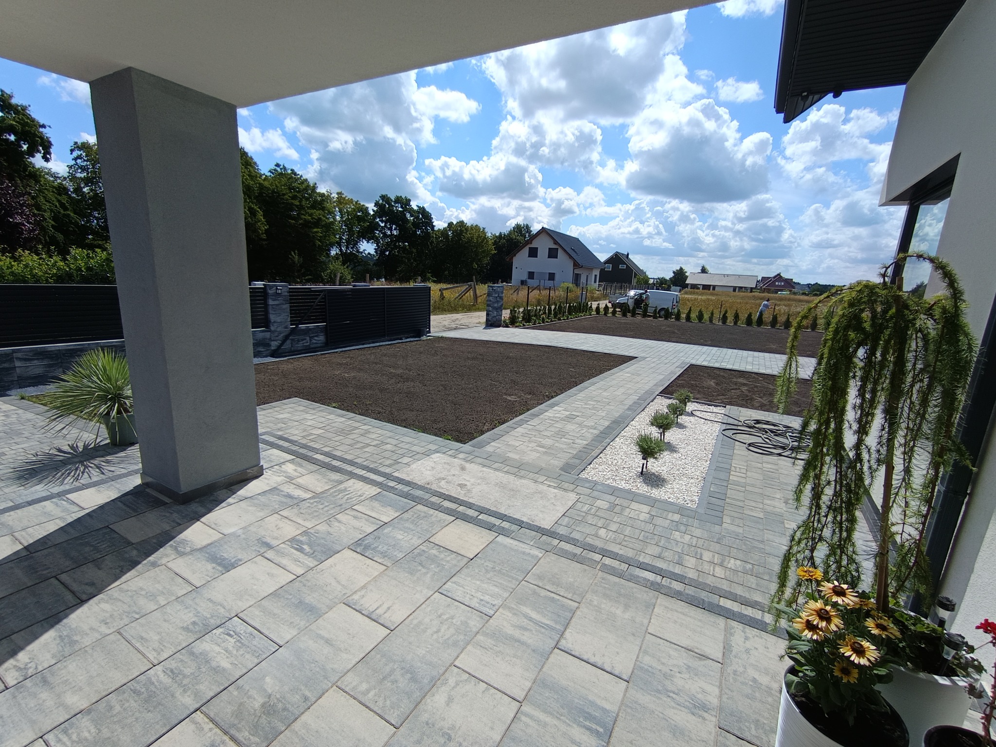 Nowoczesny taras z szarej kostki brukowej, rabaty z ziemią i białym kamieniem, w tle dom jednorodzinny i błękitne niebo z chmurami. Widok z perspektywy tarasu.