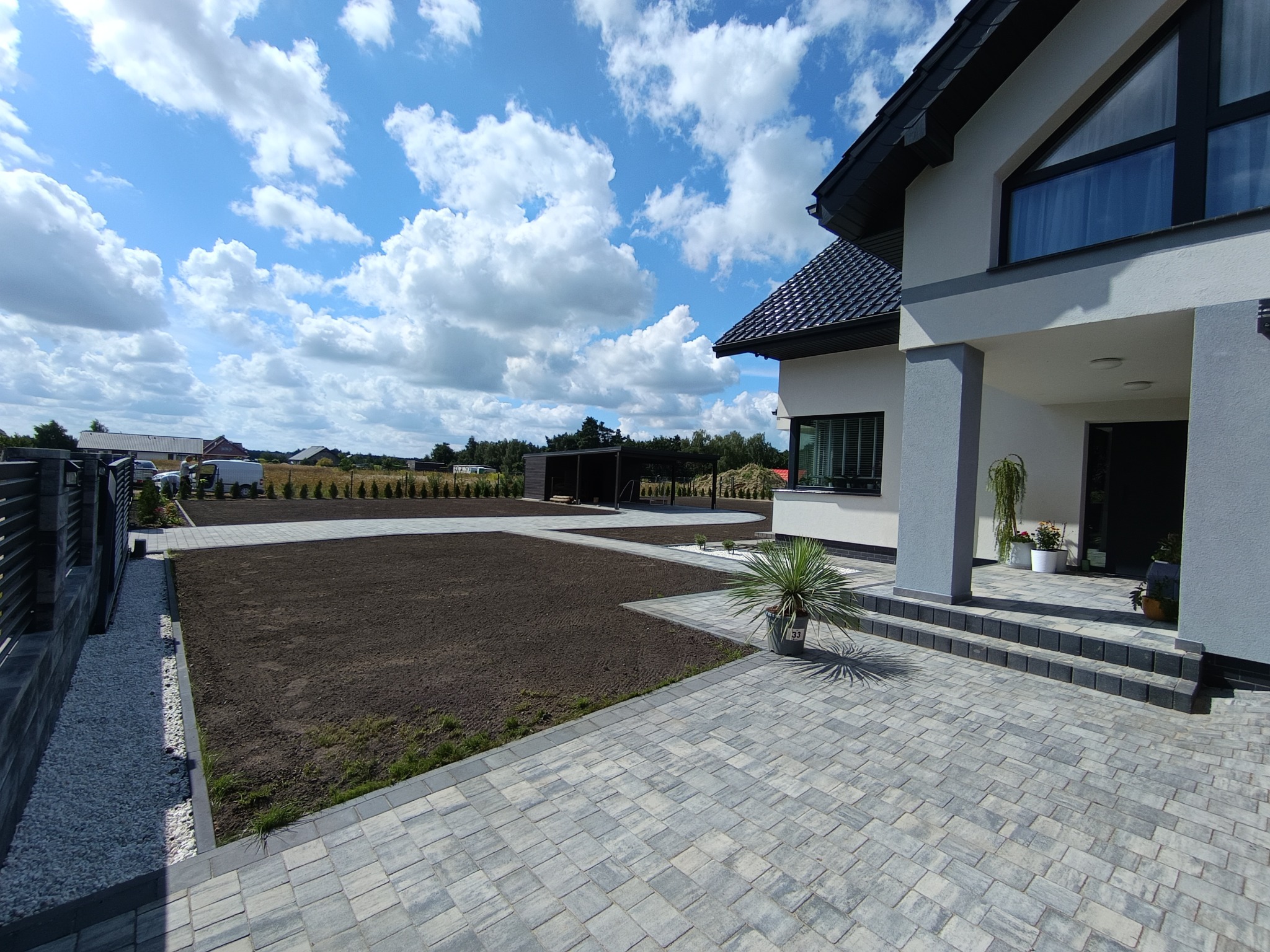 Nowoczesny dom z brukowanym podjazdem i świeżo przygotowanym terenem pod trawnik, widok z poziomu gruntu, błękitne niebo z chmurami w tle.