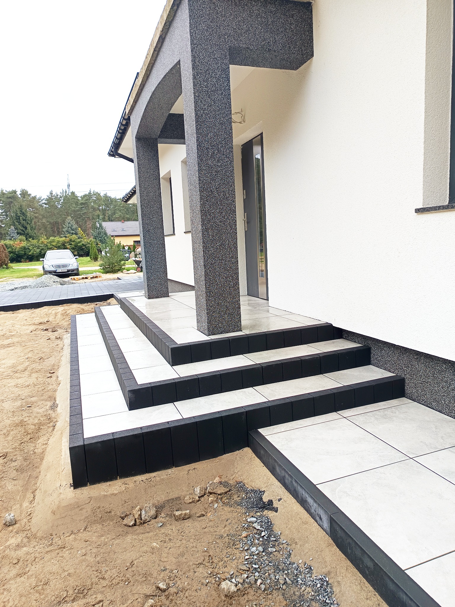 Nowoczesne schody wejściowe z białych płytek i czarnych obrzeży prowadzące do domu z szarą elewacją, widoczne z poziomu gruntu.