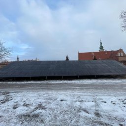 ISKRUS SPÓŁKA Z OGRANICZONĄ ODPOWIEDZIALNOŚCIĄ - Rozległa instalacja paneli słonecznych na stelażu, z widocznym budynkiem z czerwonej cegły w tle, zimowy krajobraz z resztkami śniegu na ziemi.