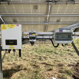 ISKRUS SPÓŁKA Z OGRANICZONĄ ODPOWIEDZIALNOŚCIĄ - Instalacja paneli słonecznych z widocznym inwerterem, zabezpieczeniami i skrzynką przyłączeniową na tle trawy. Widok od spodu konstrukcji.