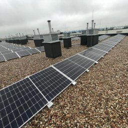 ISKRUS SPÓŁKA Z OGRANICZONĄ ODPOWIEDZIALNOŚCIĄ - Rząd paneli słonecznych na żwirowym dachu budynku z wentylacją. Widok z góry, pochmurne niebo. Instalacja fotowoltaiczna na dachu przemysłowym.