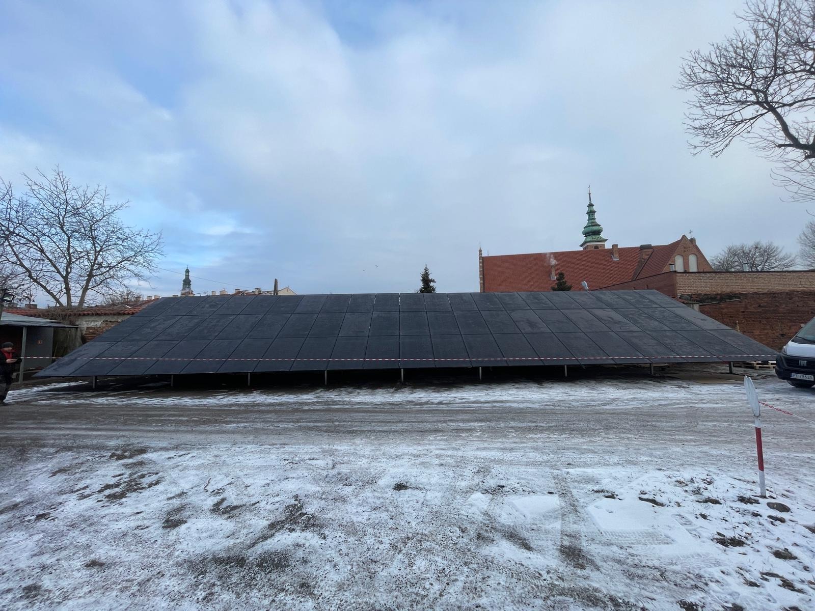 Rozległa instalacja paneli słonecznych na stelażu, z widocznym budynkiem z czerwonej cegły w tle, zimowy krajobraz z resztkami śniegu na ziemi.